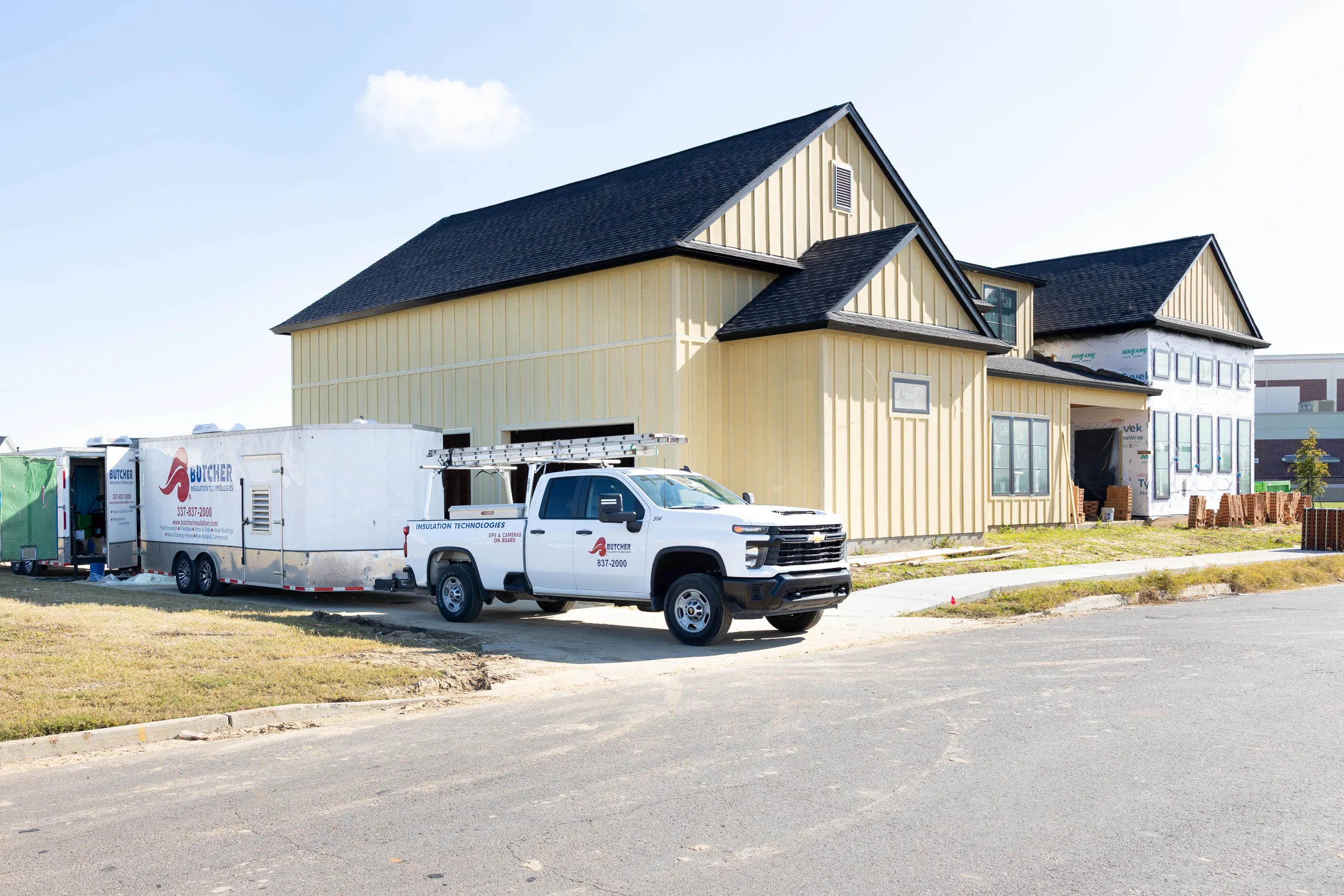 Yellow house under construction with dark roof, contractor pickup and Butcher Insulation trailers parked in driveway