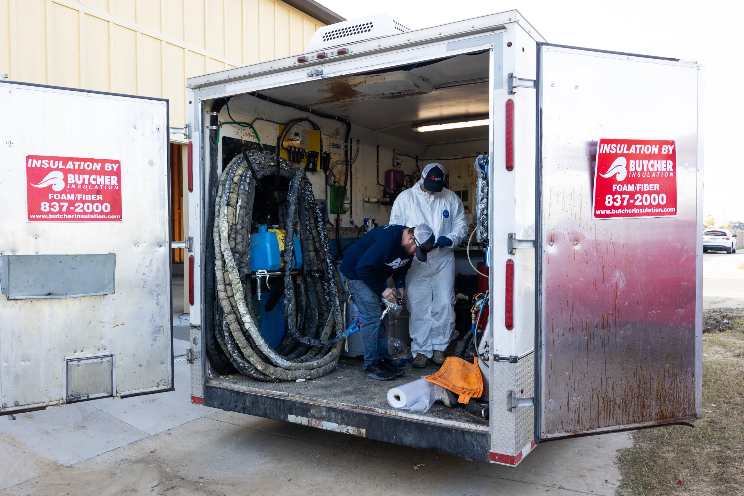 Workers prepping spray-foam equipment inside an insulation contractor's trailer with red 'Insulation by Butcher' signs.