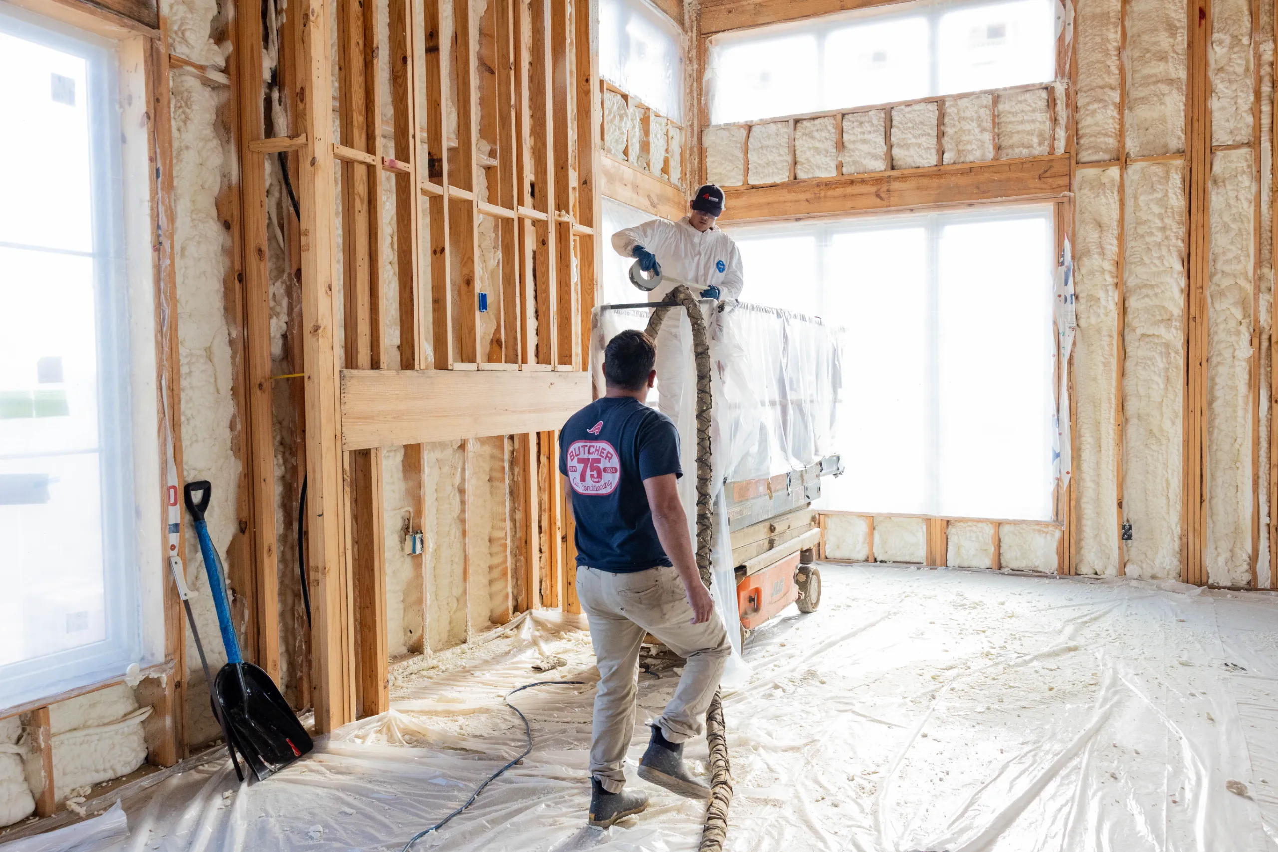 Two workers applying spray foam insulation inside framed home, protective suits and equipment, wooden stud walls.