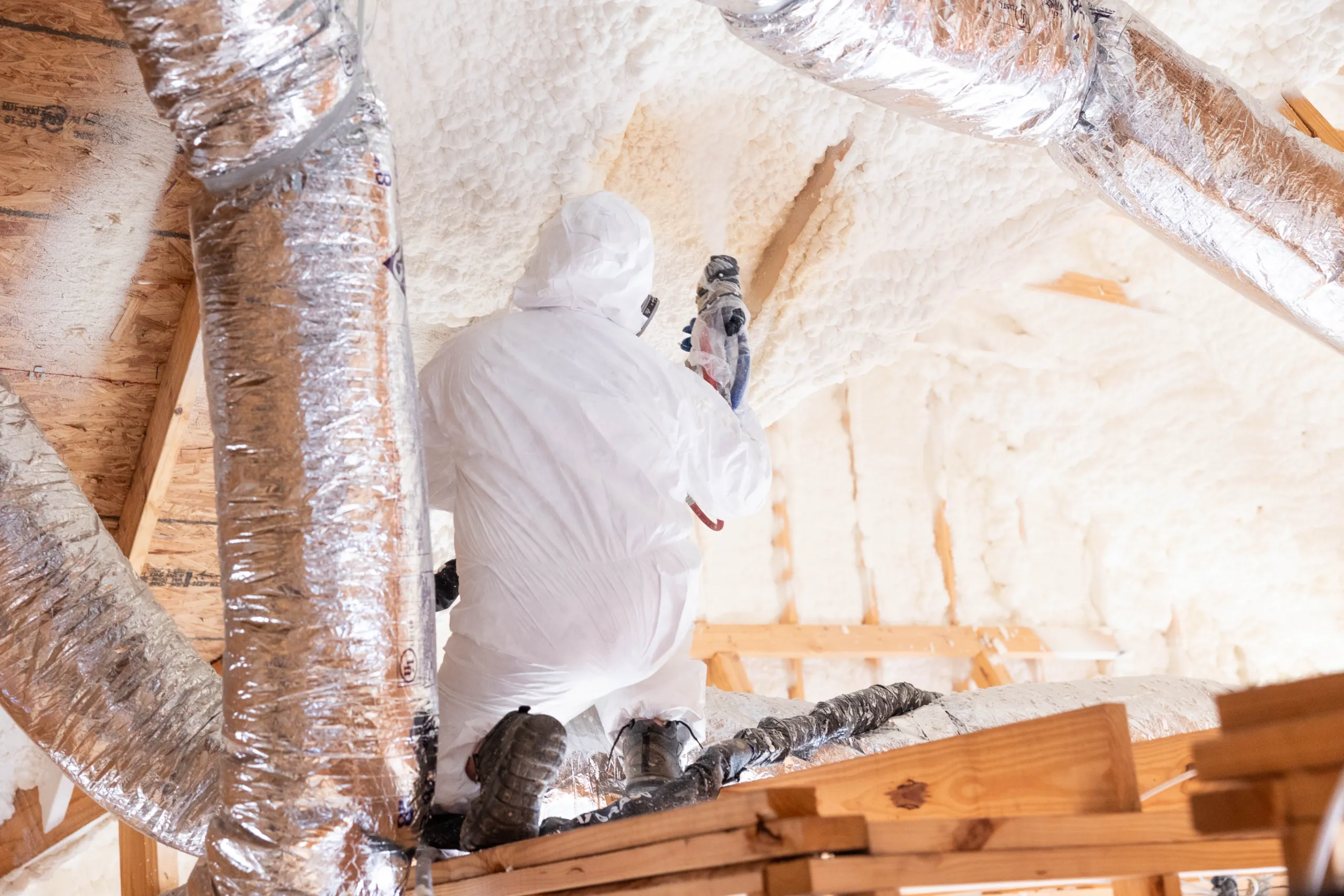 A person in a protective suit applying spray foam insulation to an attic ceiling near HVAC ductwork