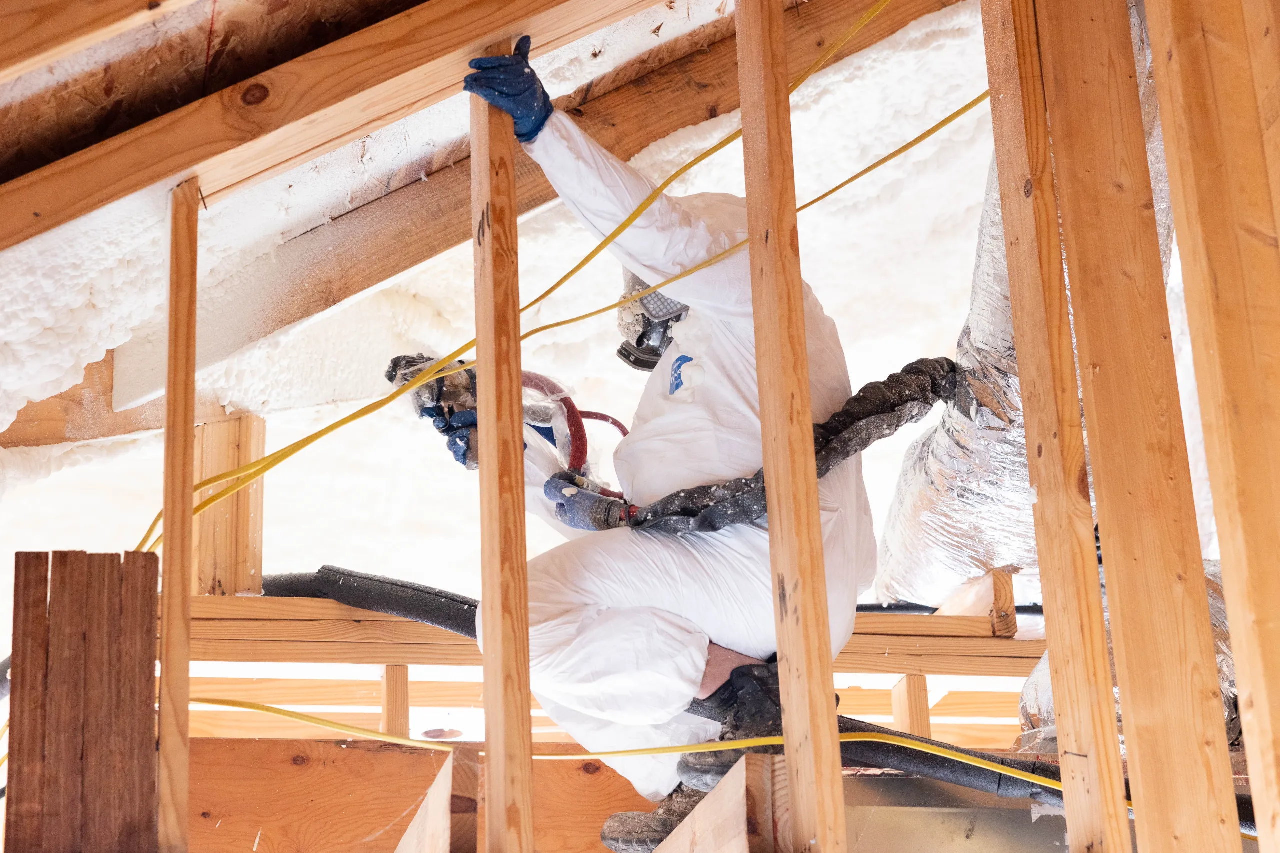 A worker in a protective suit applying spray foam insulation in an attic between wooden studs