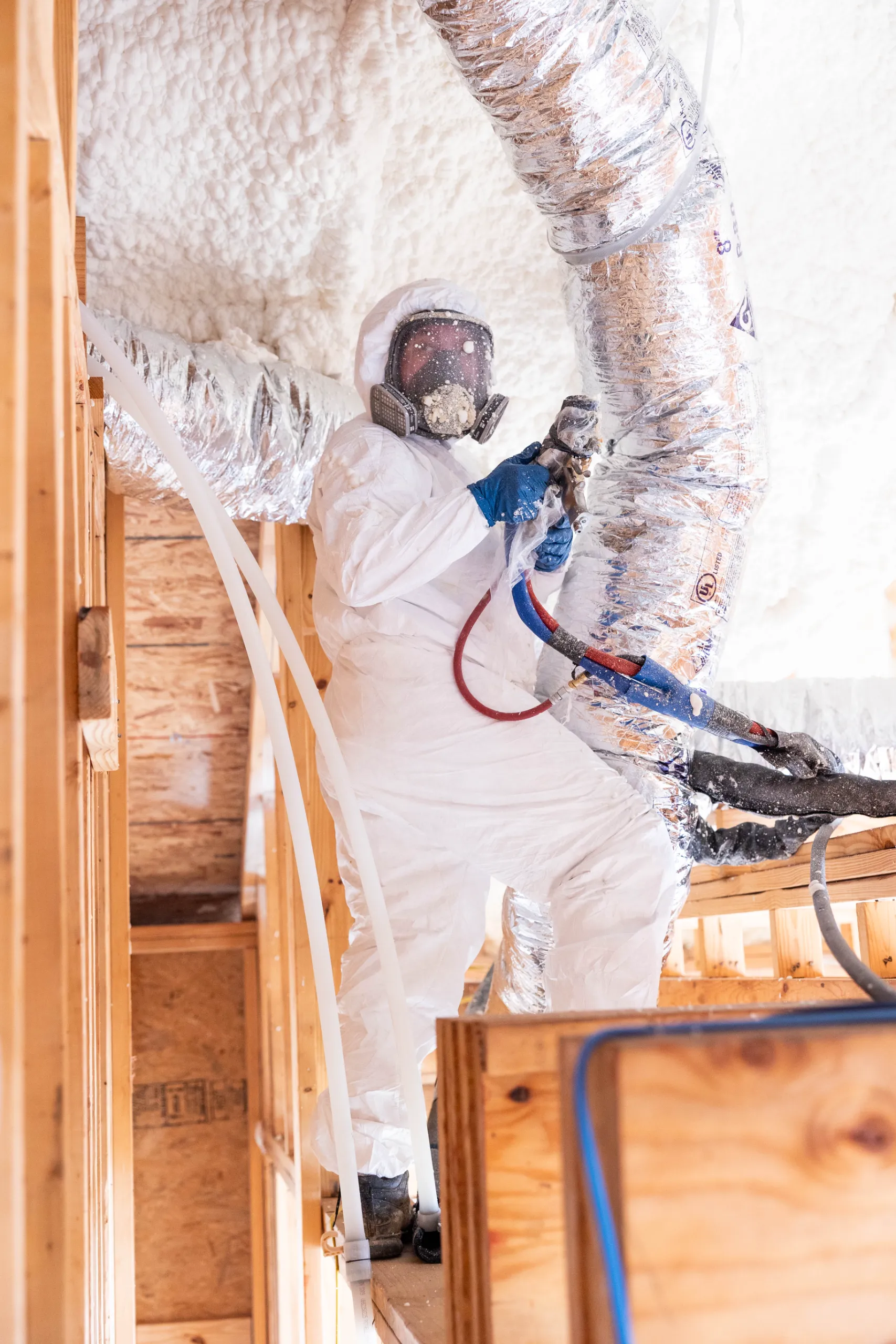 A worker in protective suit and respirator spraying spray foam insulation around attic ductwork