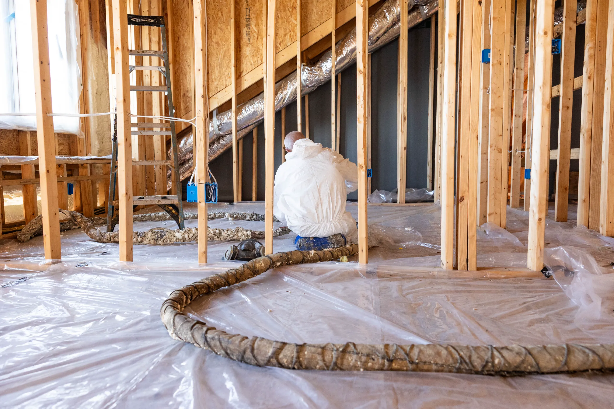 A worker in a white protective suit applying spray foam insulation in a framed attic with plastic sheeting and ductwork