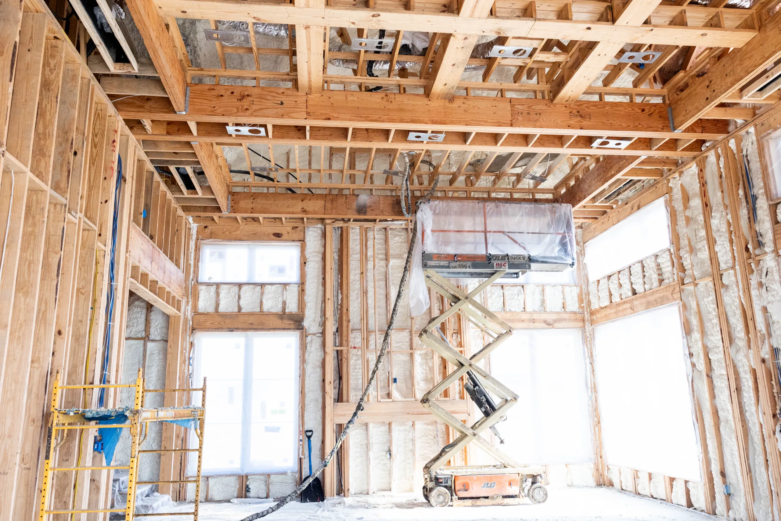 Residential interior under construction with exposed wood framing, spray foam insulation, scaffolding and scissor lift
