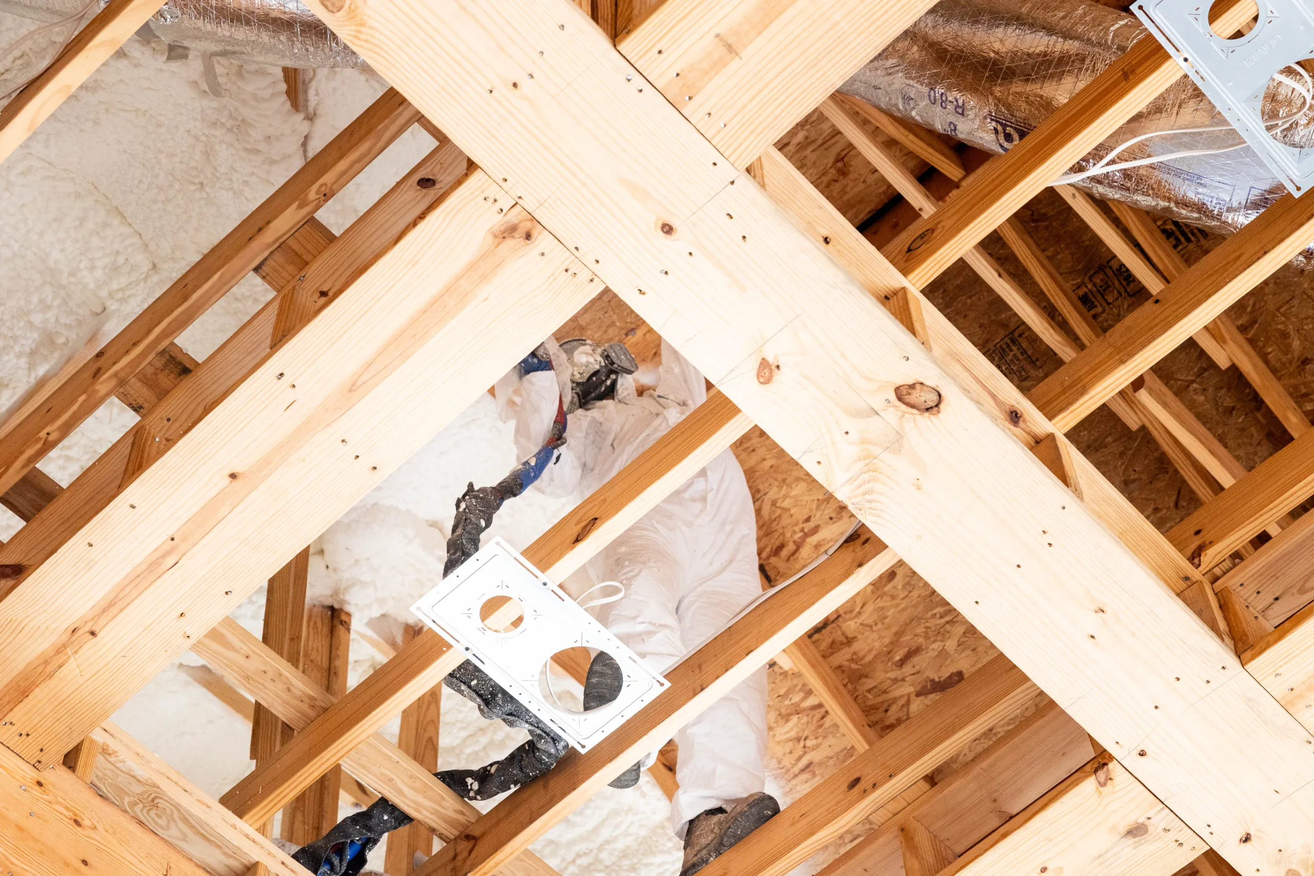 A worker in protective suit installing spray foam insulation in an attic framed by wooden roof trusses