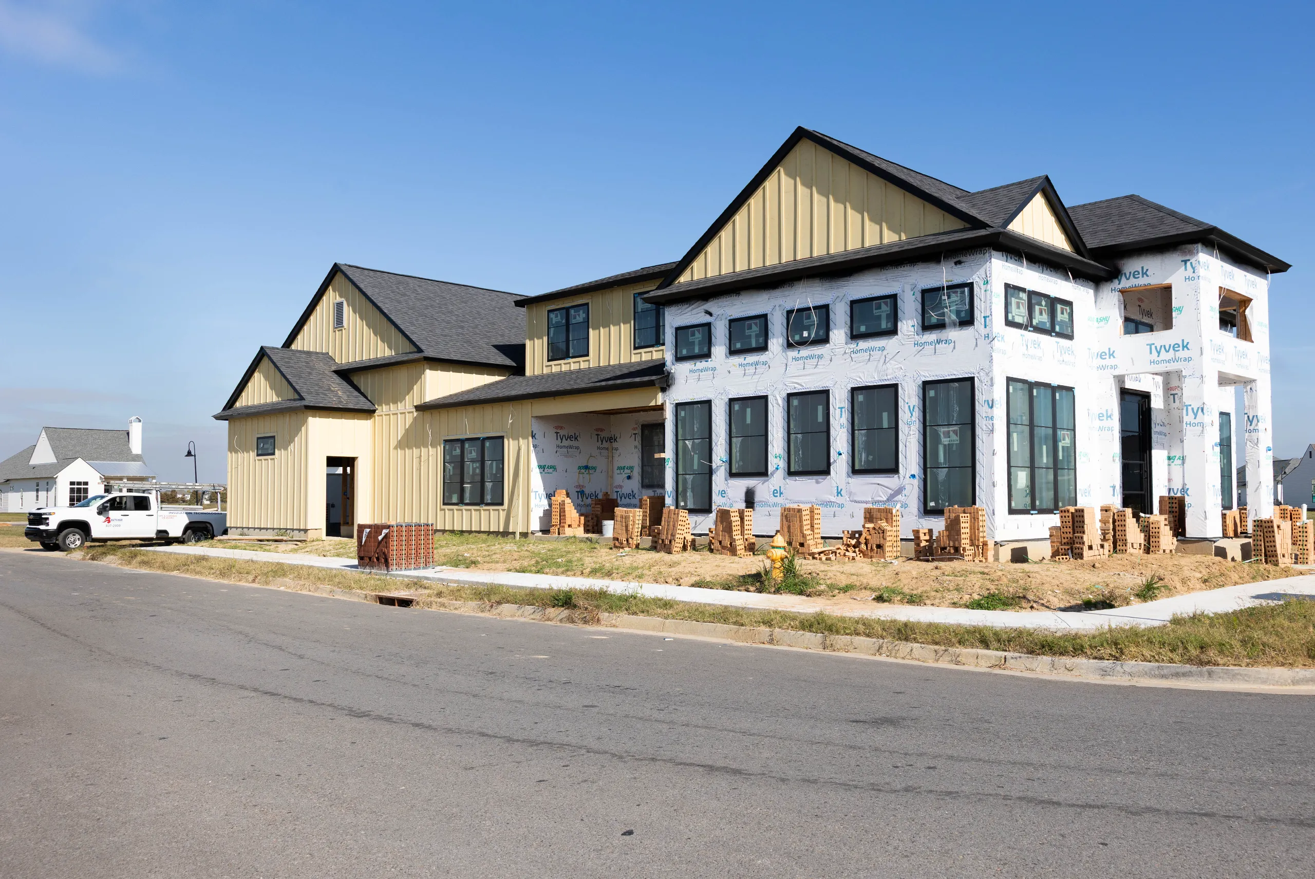 Large modern farmhouse-style house under construction with yellow siding, Tyvek wrap, stacked bricks and a white work truck.