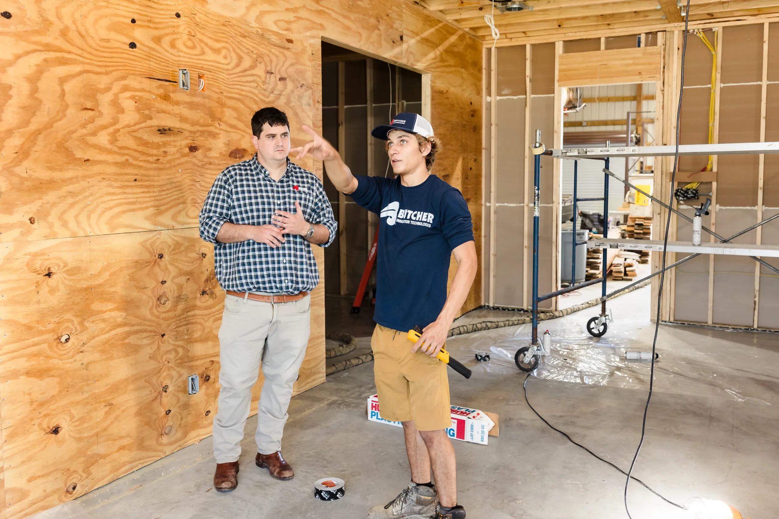Construction worker pointing as a man in a plaid shirt listens inside a plywood-framed building