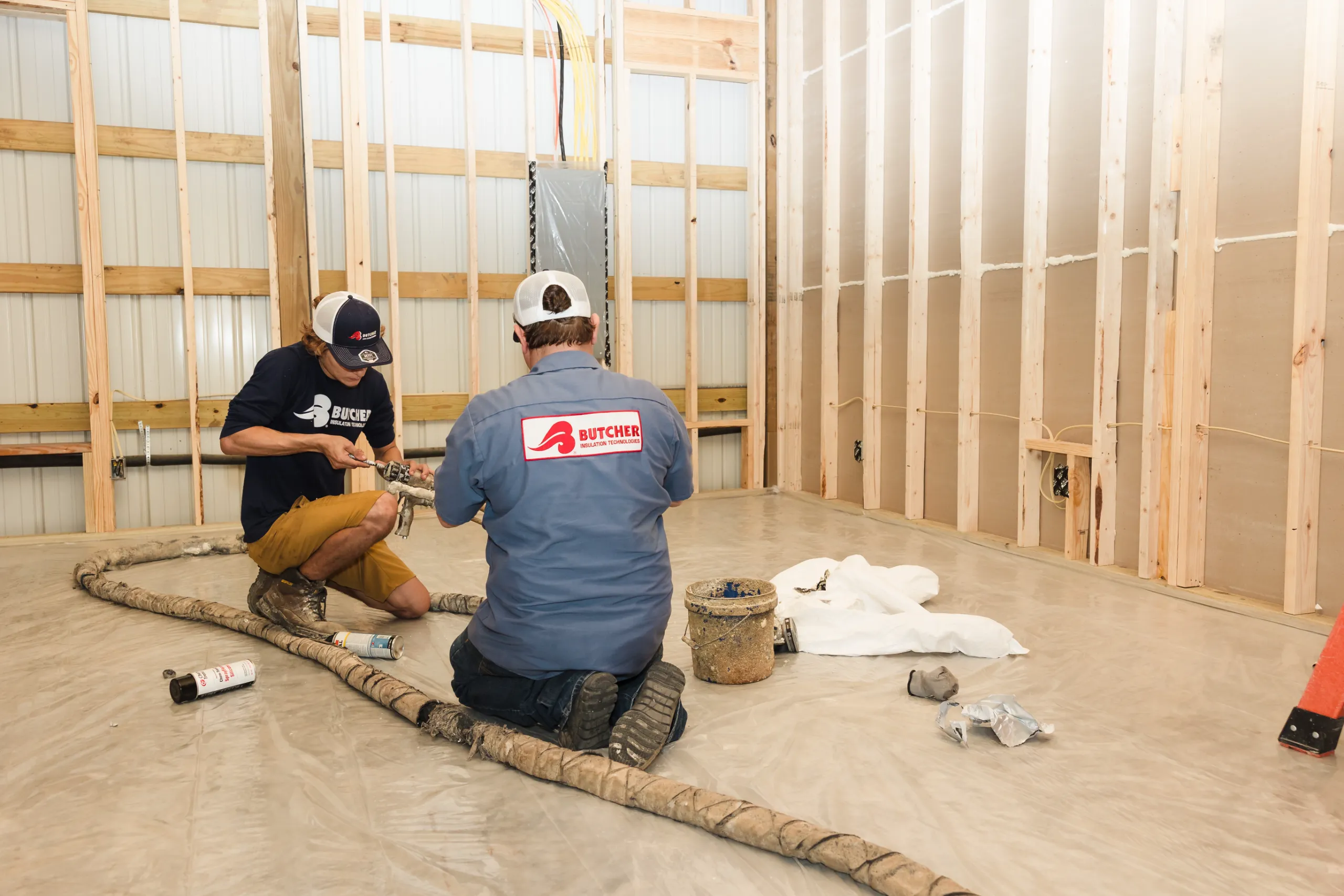 Two workers kneeling and prepping spray foam insulation hose inside unfinished framed room with exposed wooden studs.