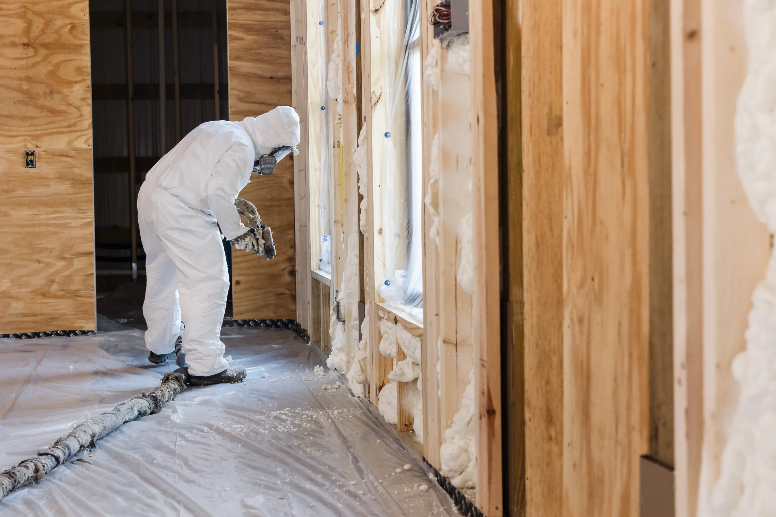 A person in a white protective suit spraying foam insulation into wooden wall studs on a construction site