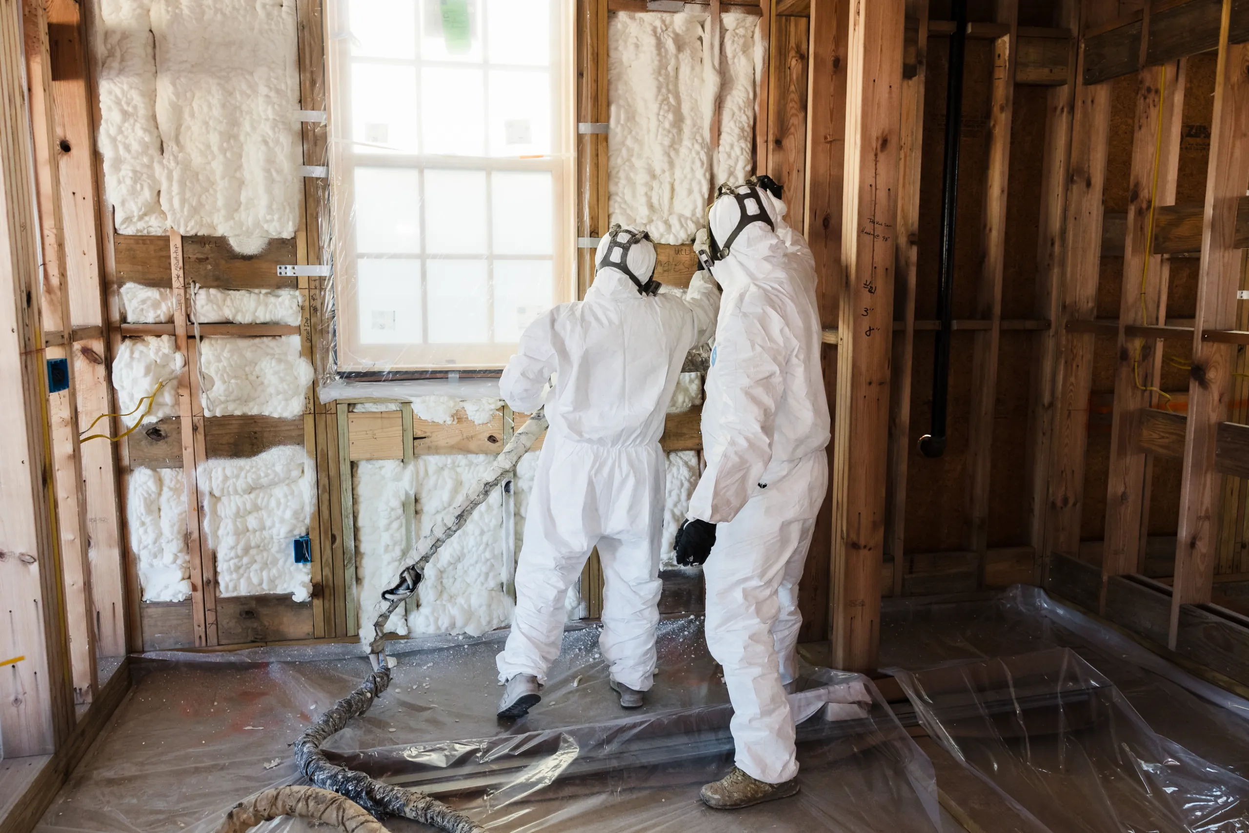 Two workers in white protective suits spraying spray-foam insulation into studs around a window in a framed room