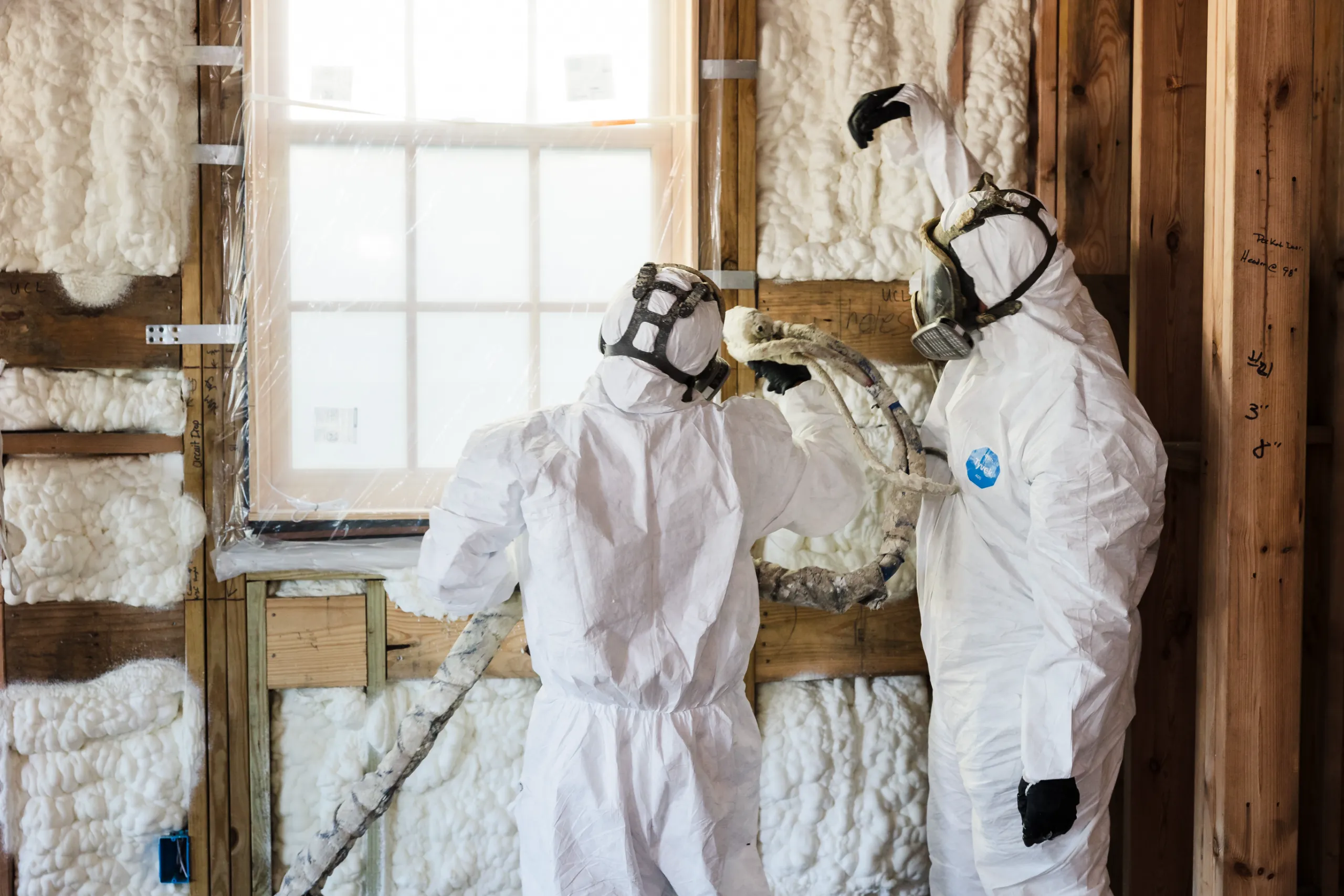 Two workers in protective suits and respirators applying spray foam insulation to a wood-framed wall