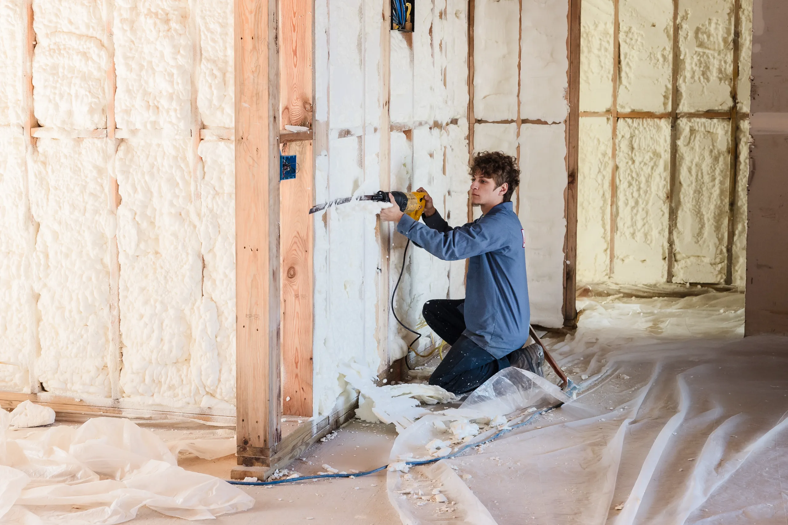 A man kneeling installs spray foam insulation in a wall cavity using a reciprocating saw during home renovation