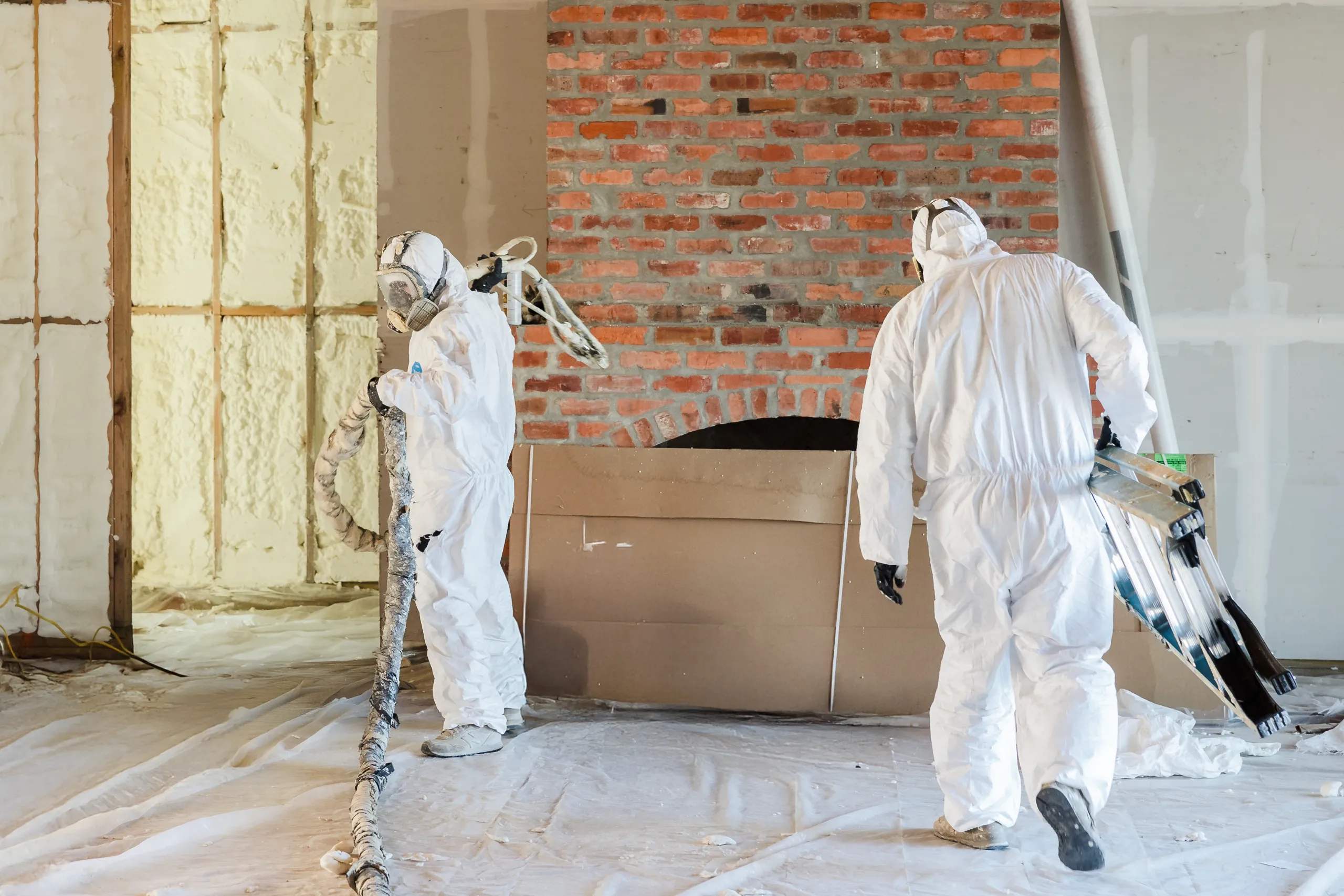 Two people in white protective suits spray foam insulation inside a house by a brick wall.