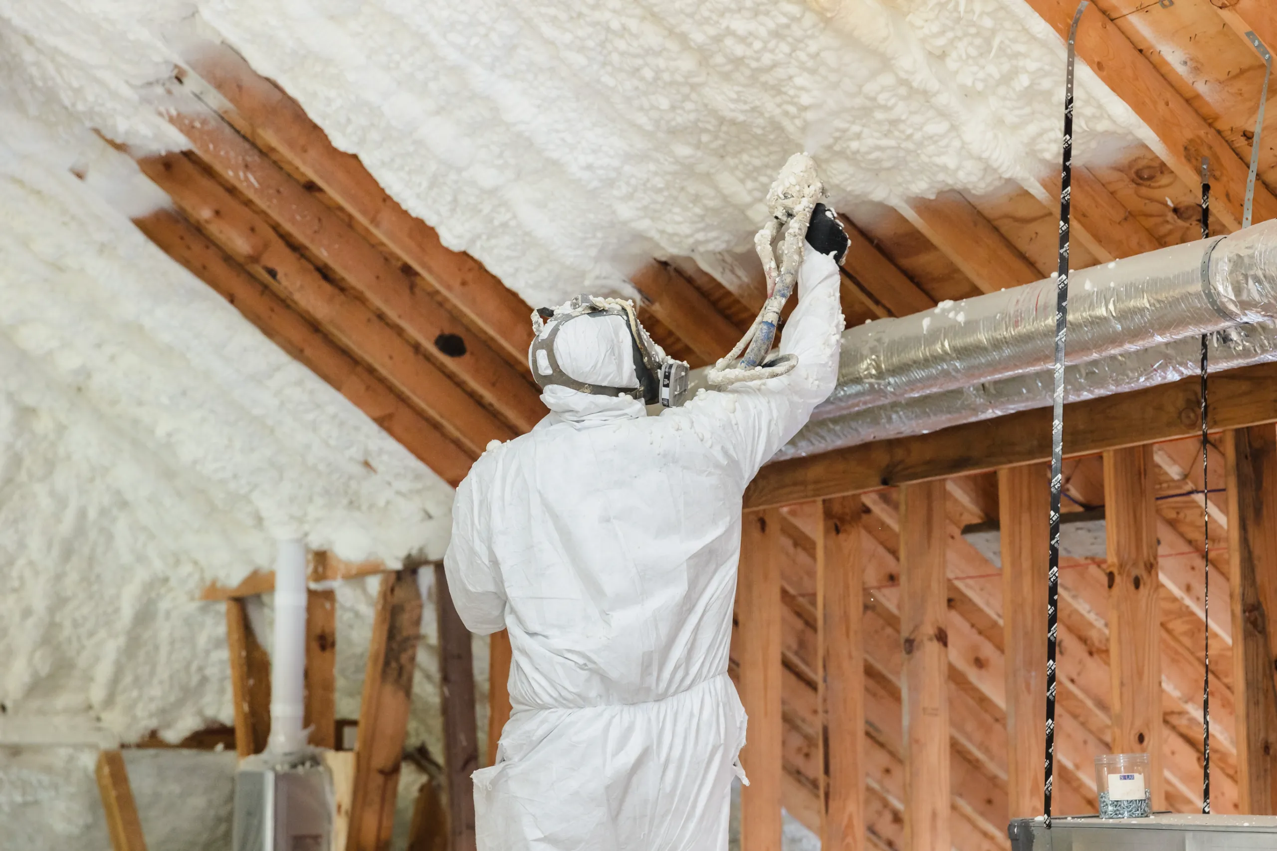A worker in protective suit spraying spray foam insulation onto attic rafters with a spray gun