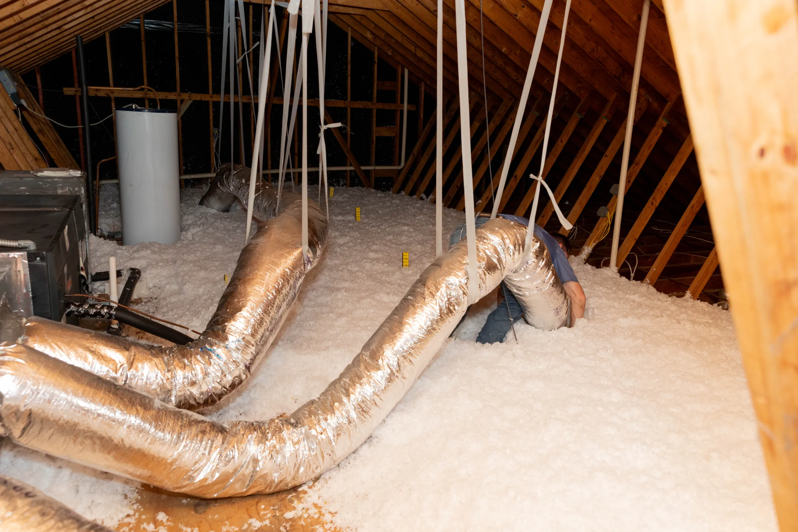 A person installing flexible HVAC ducts in an attic with blown-in insulation and exposed wooden rafters.