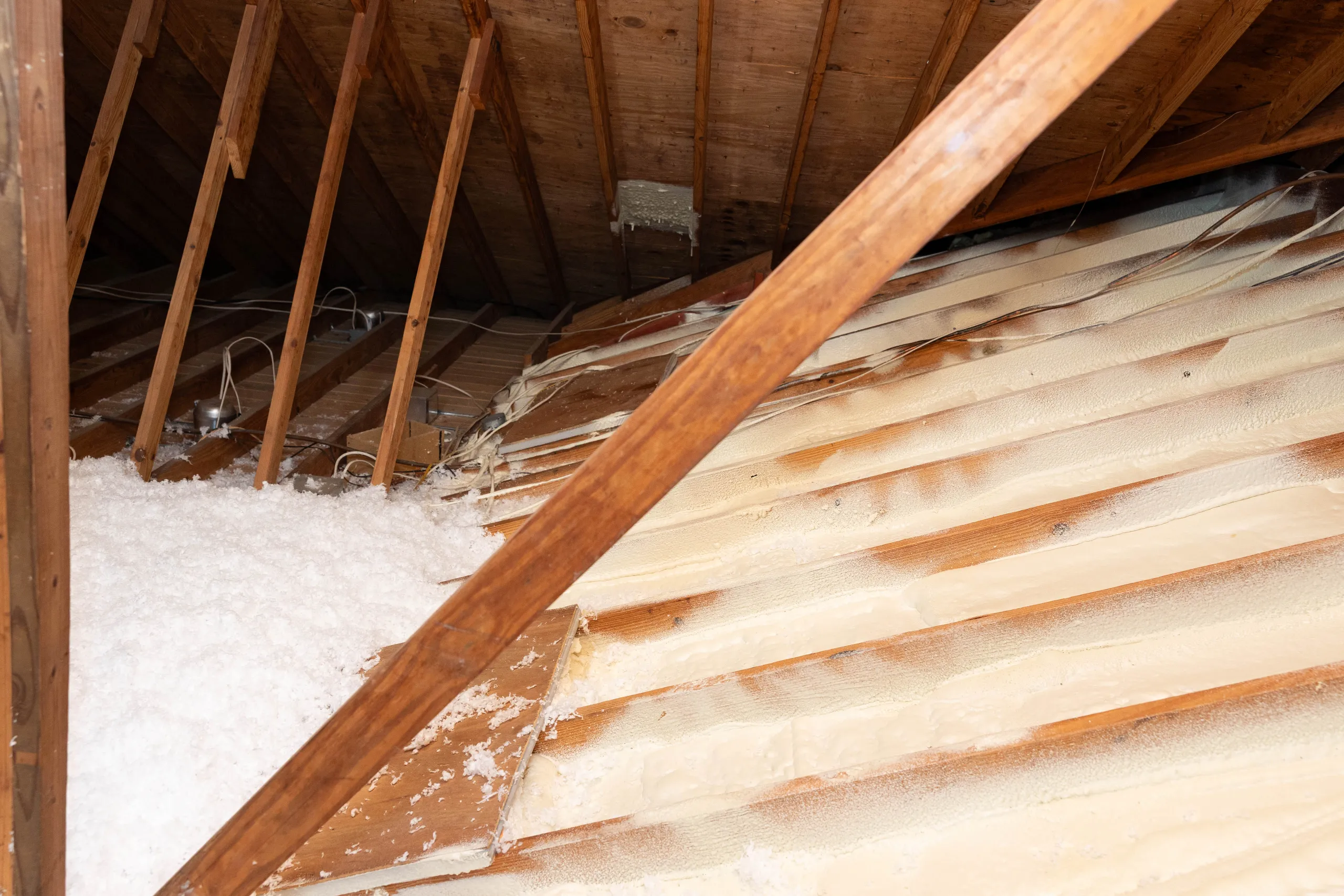 Attic with exposed wooden rafters, blown-in loose-fill insulation and spray-foam insulation on roof deck with wiring.