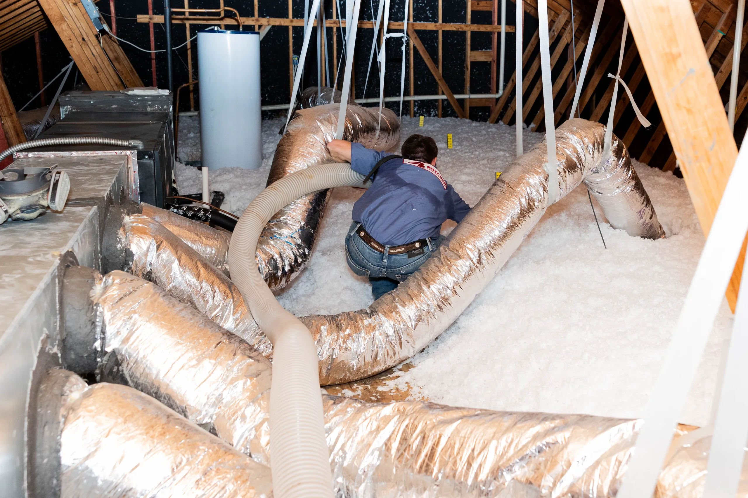 A person installing blown-in attic insulation with a hose amid foil-wrapped HVAC ducts in an attic.