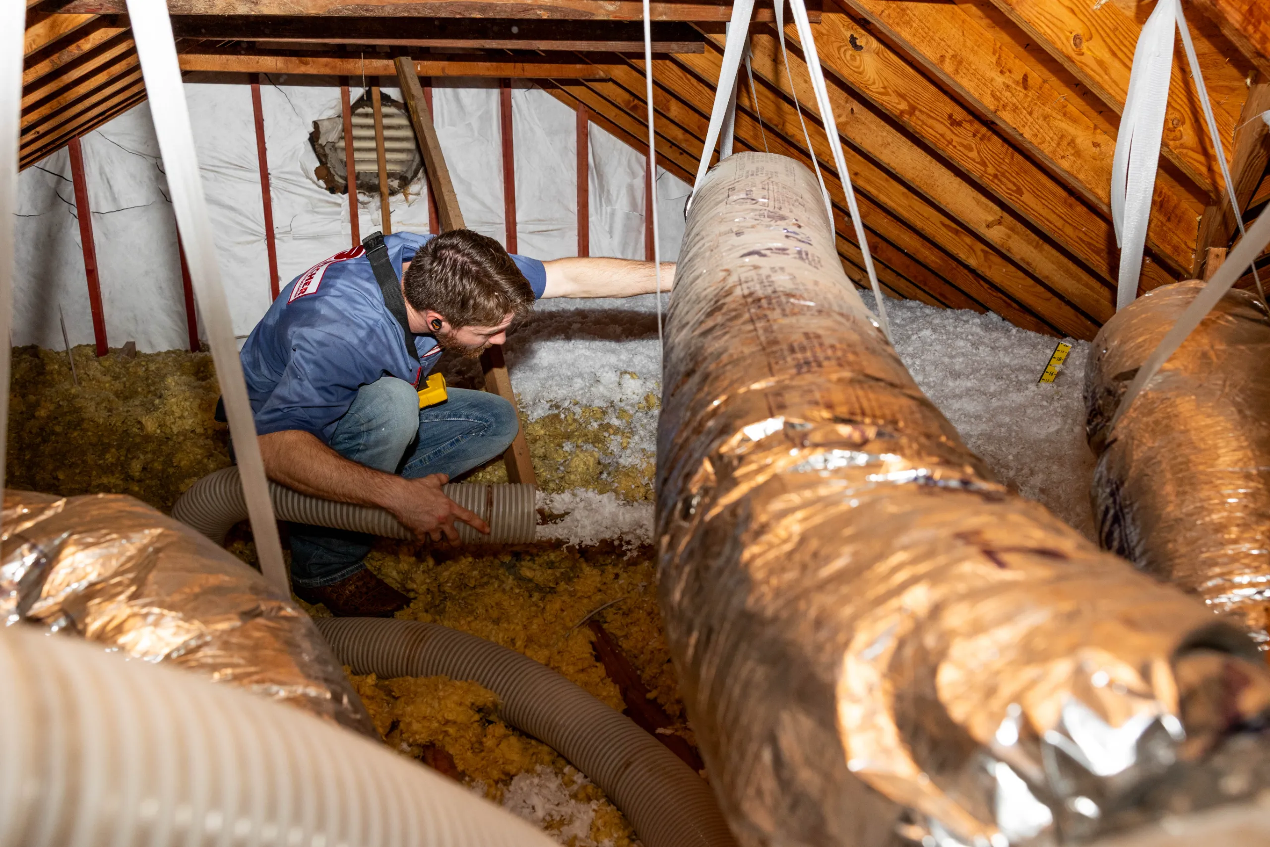 A man installing blown-in attic insulation, guiding a flexible hose around HVAC ducts and wooden joists.
