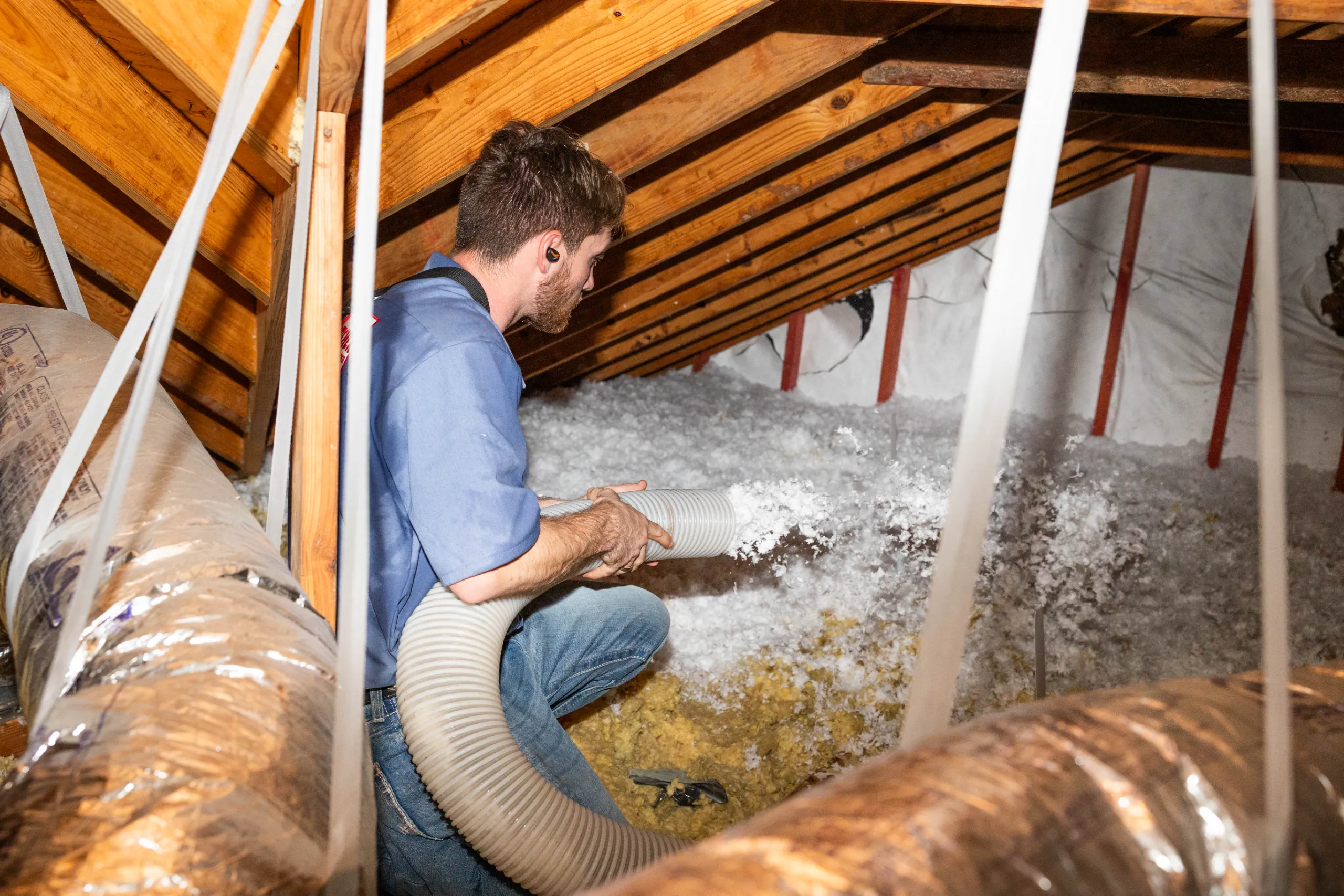 A man installing blown-in attic insulation with a large flexible hose under wooden roof rafters