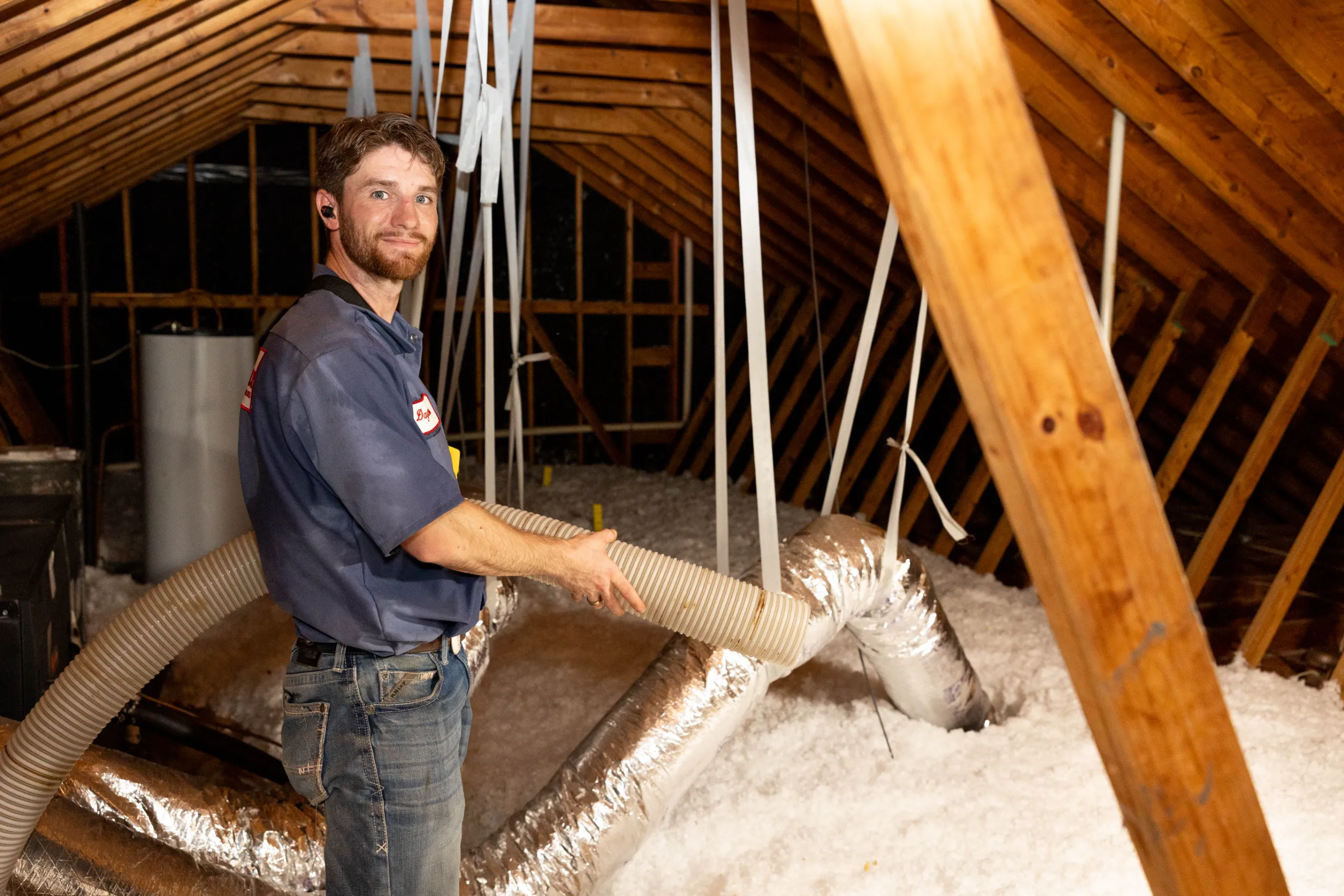 A man installing blown-in attic insulation, holding a large flexible hose between wooden rafters