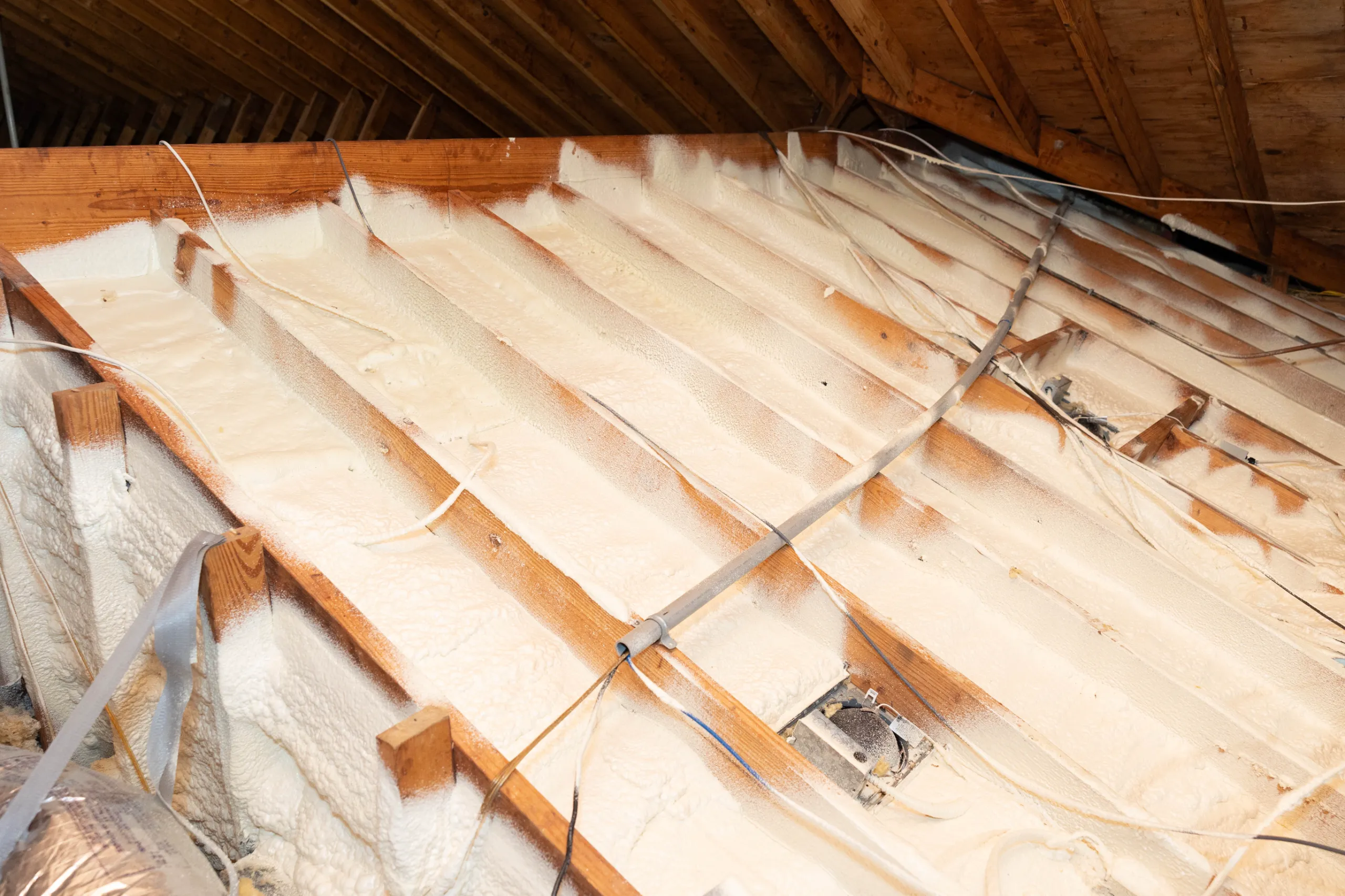 Attic with spray foam insulation between wooden rafters and exposed wiring and ductwork