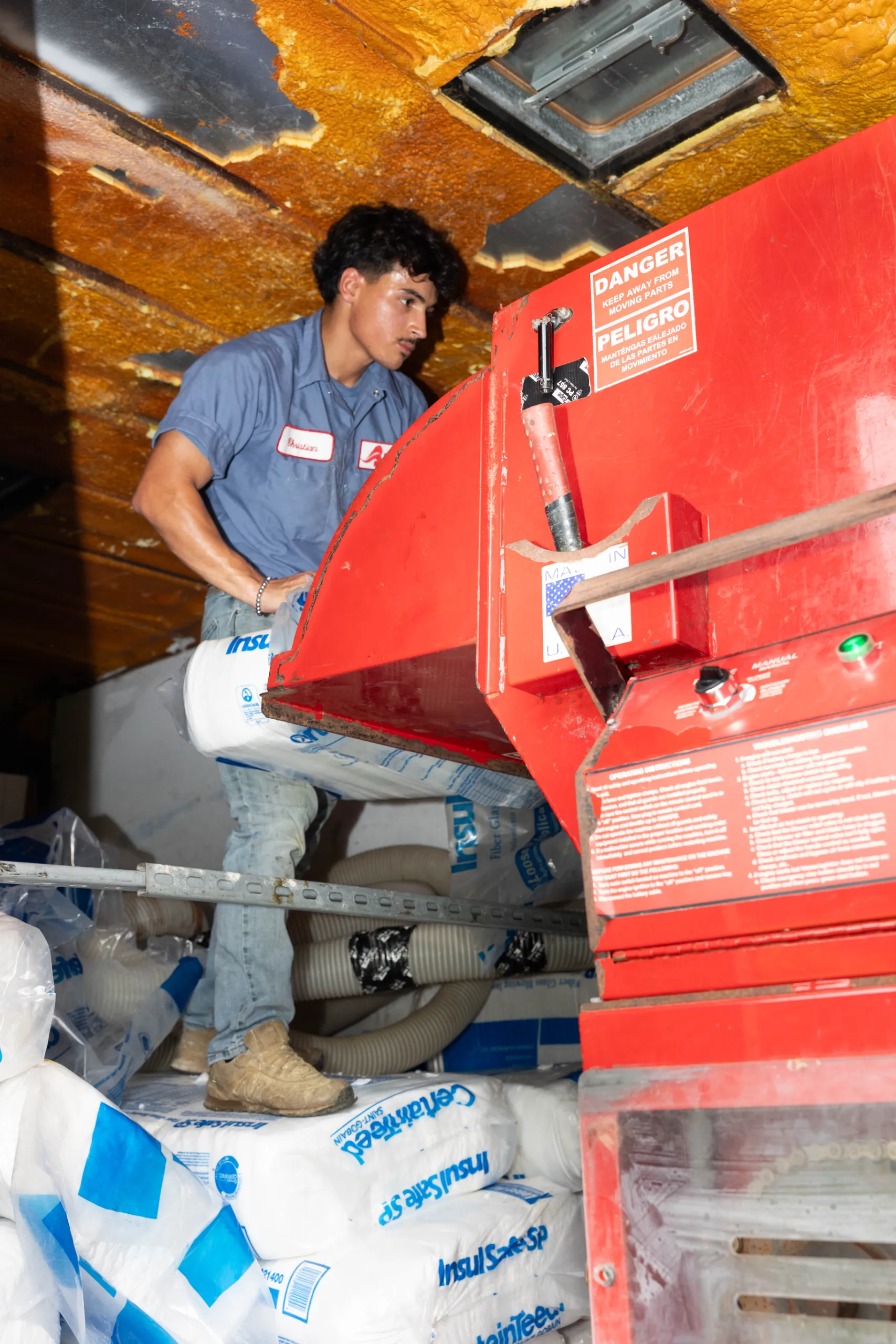 A worker installing attic insulation, handling insulation bags beside a large red HVAC machine with danger label
