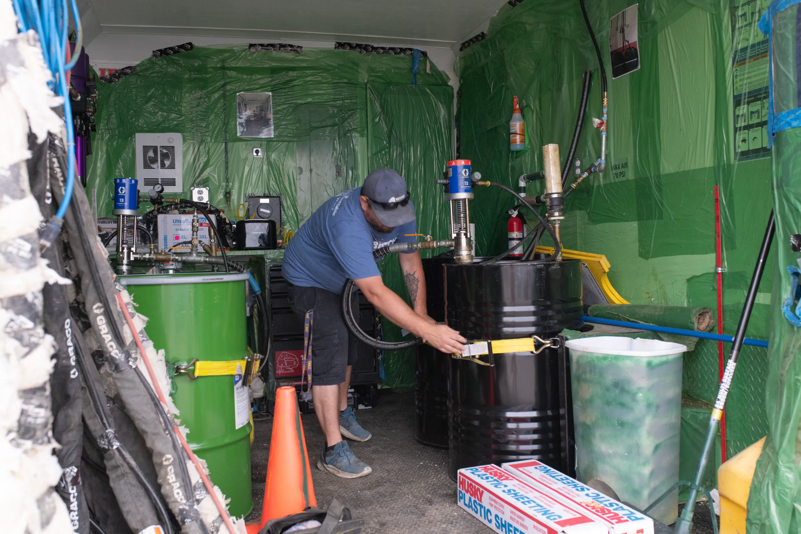 A man operating hoses to pump liquid into black drums inside a green-lined mobile work van with safety cones.