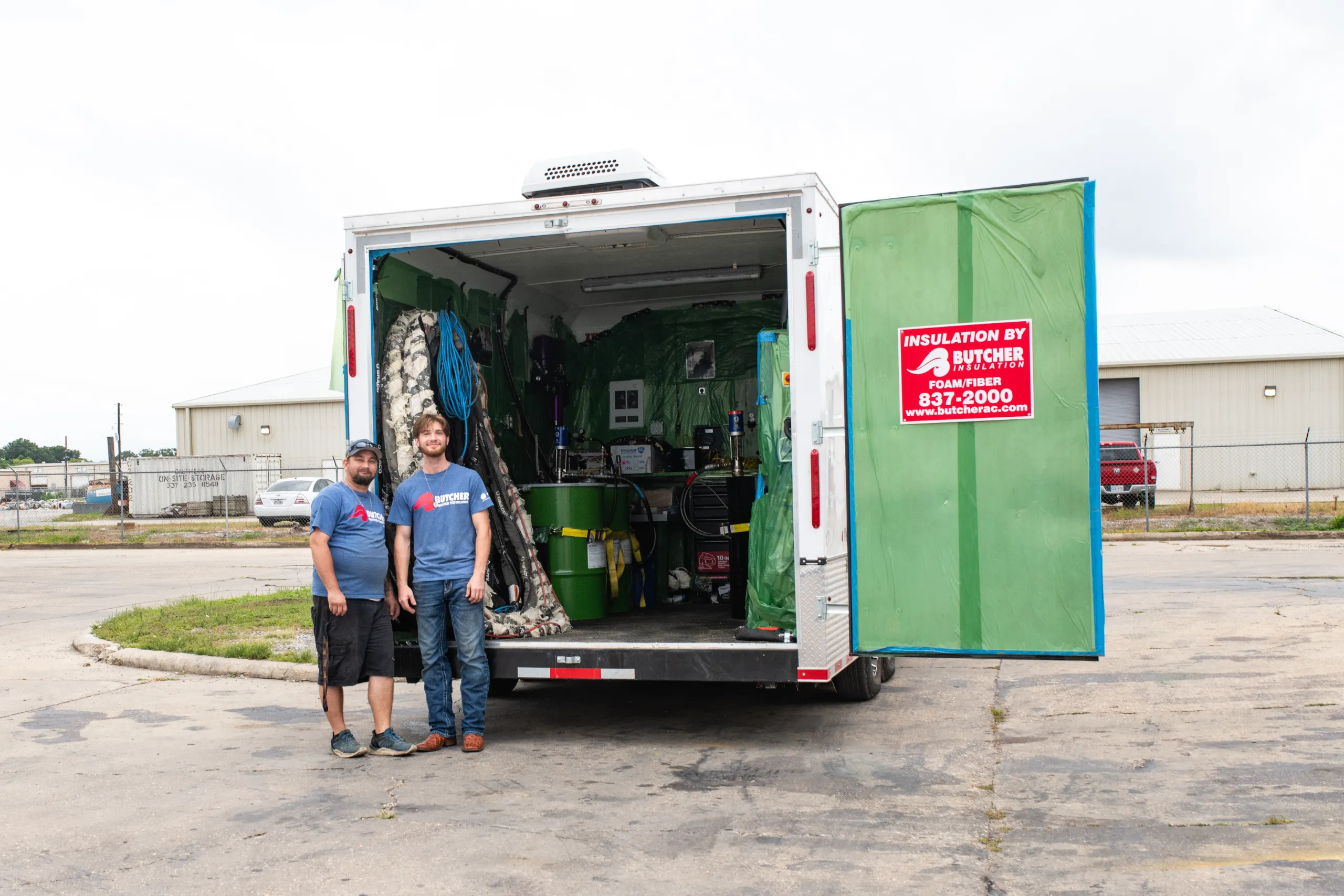 Two men in blue shirts standing by open trailer with insulation equipment and green tarp, red Butcher Insulation sign
