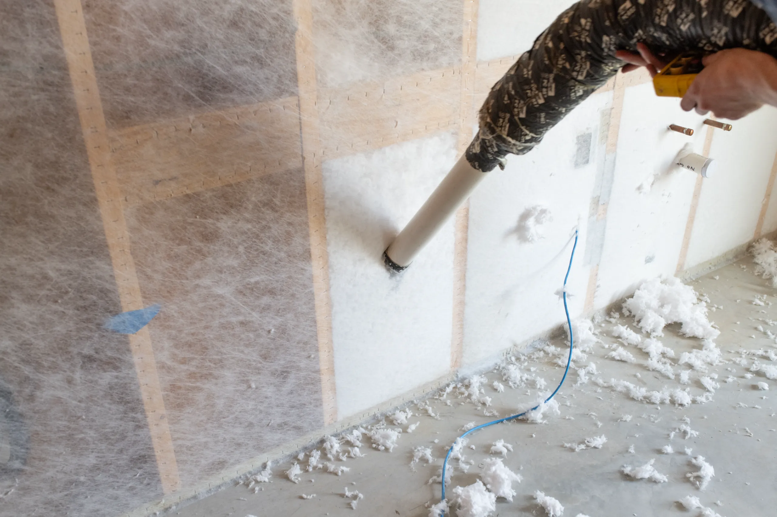 A person blowing loose-fill cellulose insulation into wall cavities with hose, scattered insulation on floor