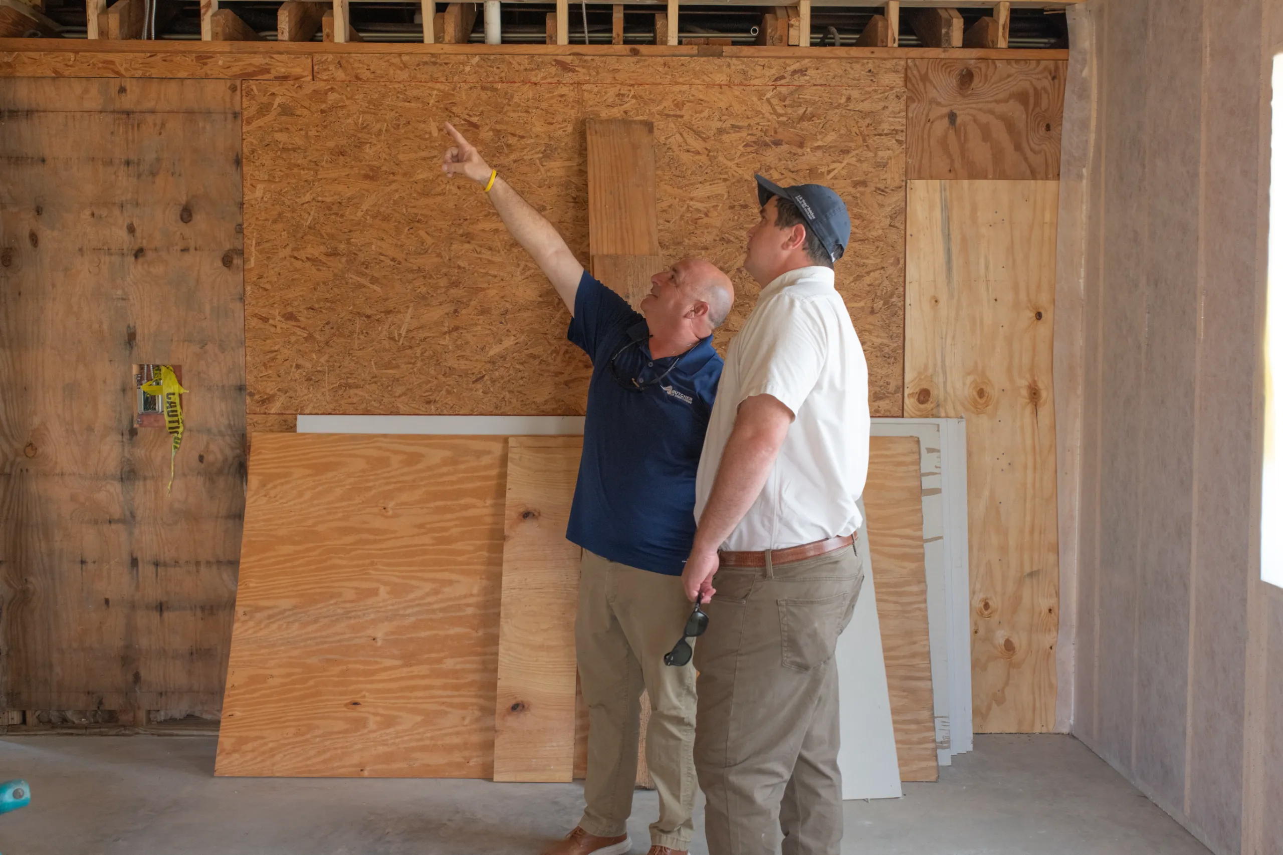 Two men inspecting plywood wall during home renovation, one pointing upward
