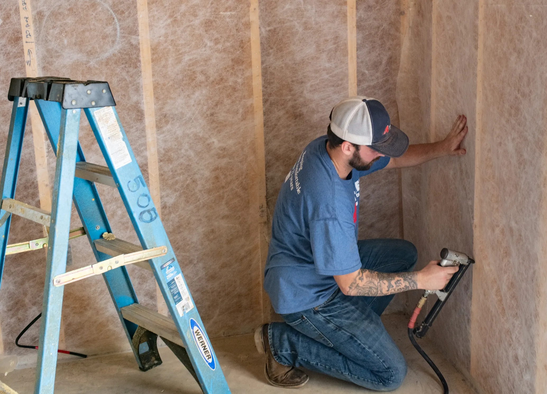 A man installing wall insulation with a nail gun beside a blue ladder in a construction interior