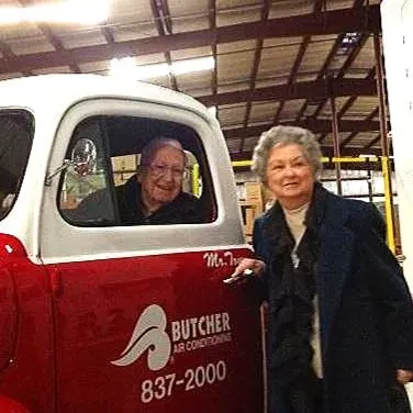 Elderly man in red Butcher Air Conditioning truck and elderly woman standing at door, smiling in warehouse