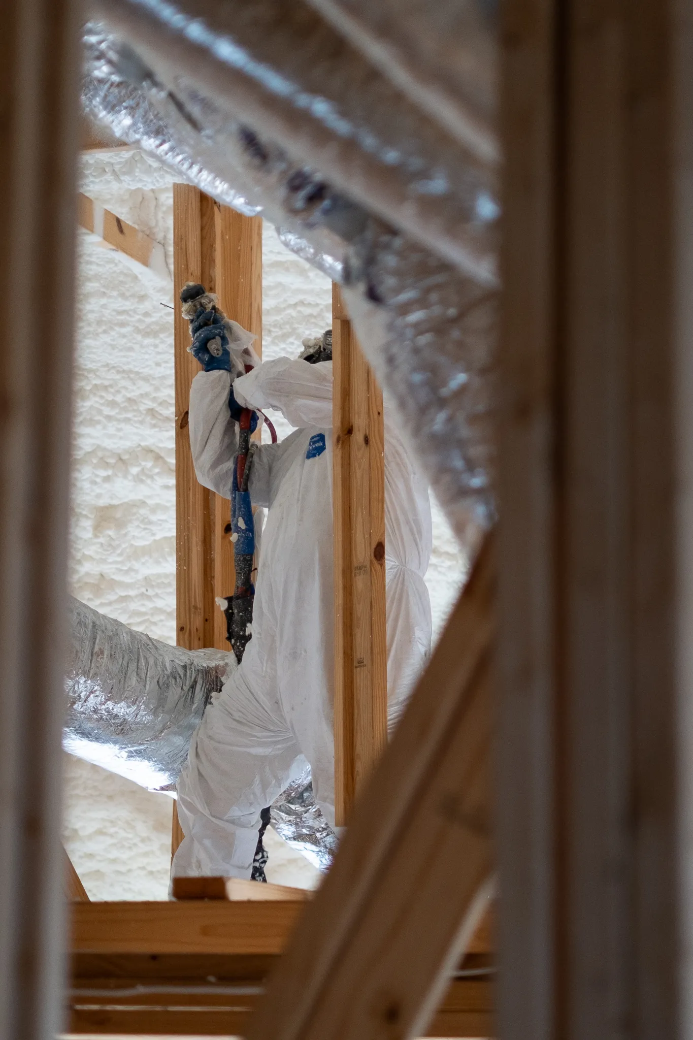 A worker in a white protective suit spraying spray foam insulation into attic wall cavities between wooden studs