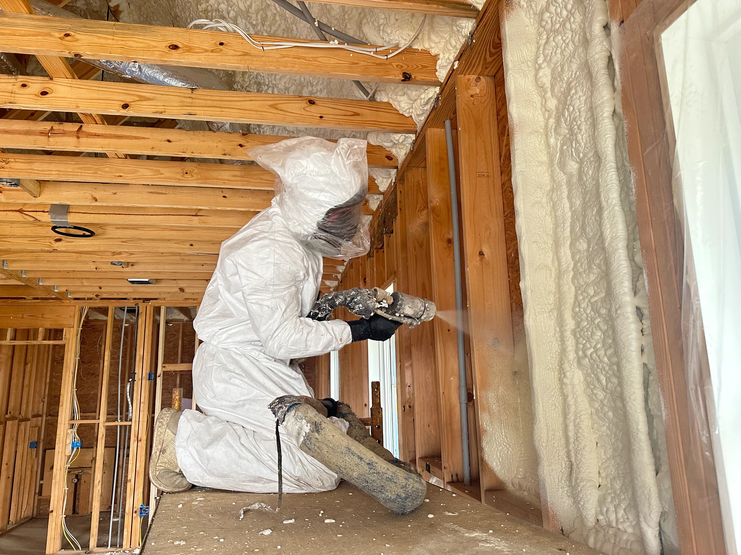 A person in a white protective suit spraying spray foam insulation onto wooden wall studs in a framed building