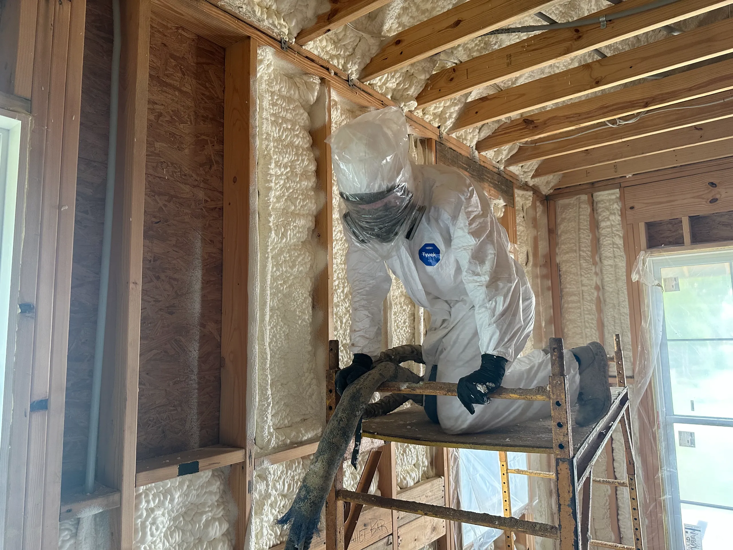 A person in protective suit applying spray foam insulation to wood-framed walls and ceiling