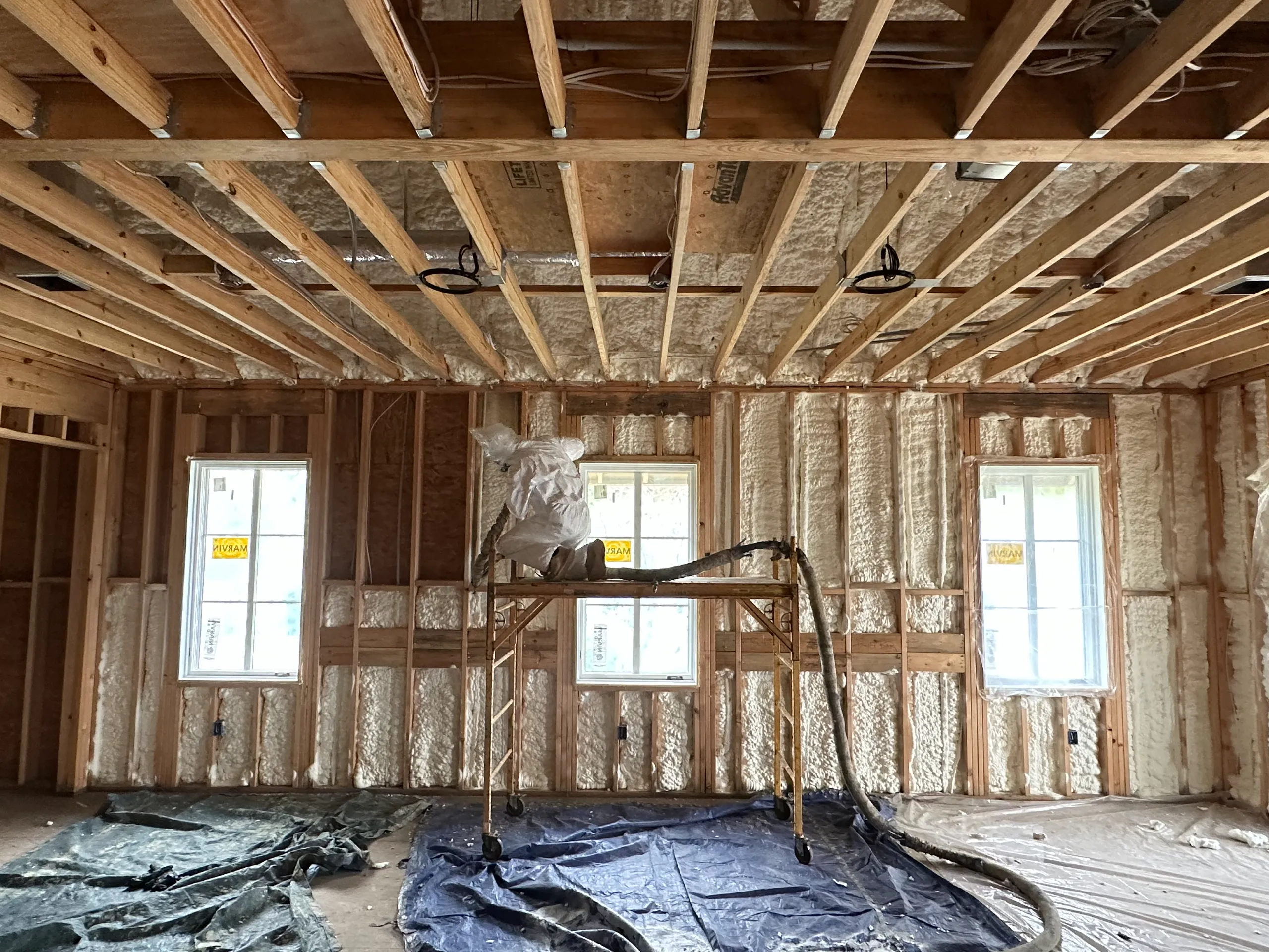 Worker in protective suit spraying spray foam insulation in unfinished framed room with exposed ceiling joists and windows