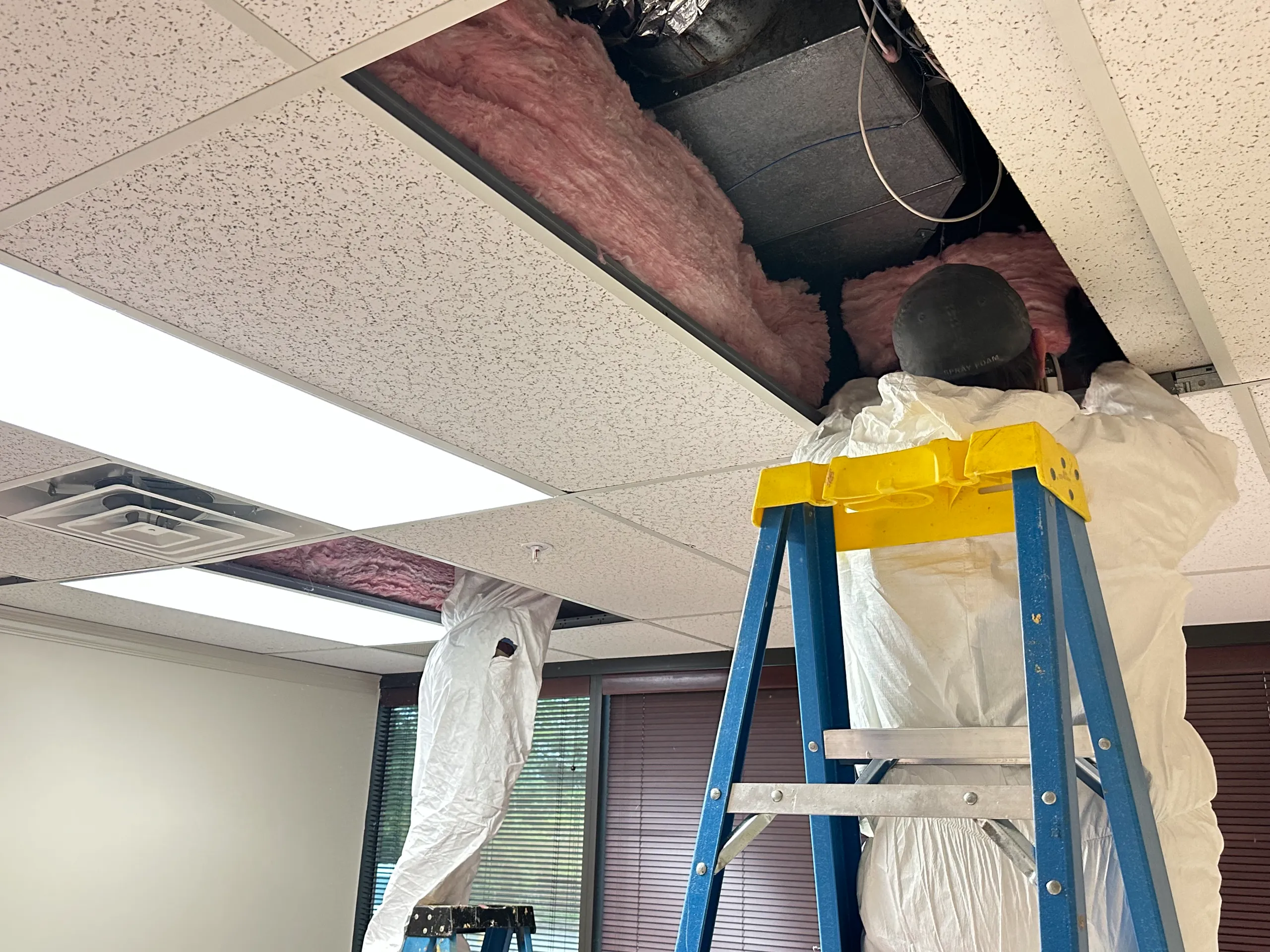 A person in protective suit on ladder installing pink fiberglass insulation into an office drop ceiling
