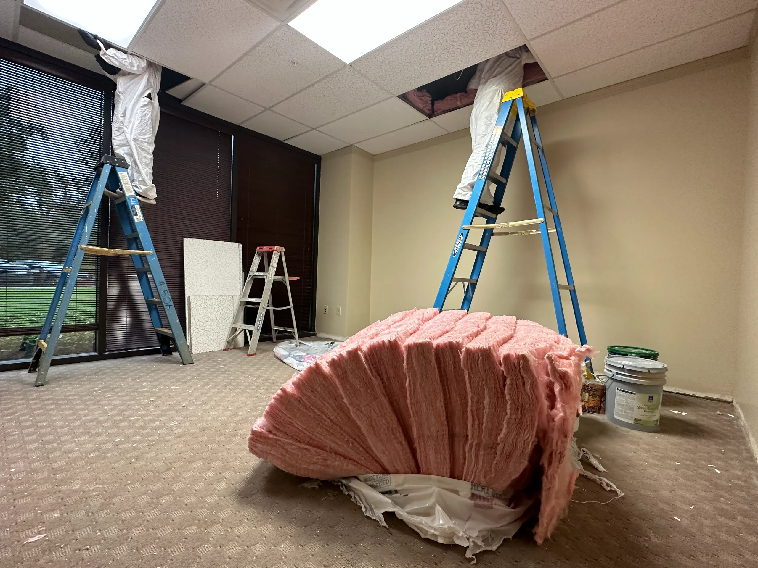 Two workers in white coveralls on ladders installing pink insulation through office ceiling tiles
