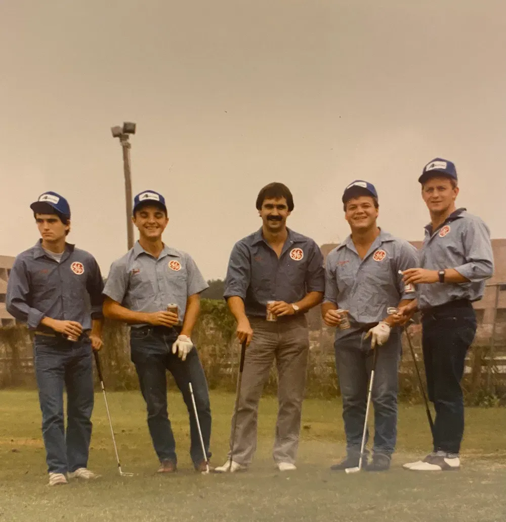 Vintage photo of five men in matching blue shirts and caps holding golf clubs and drinks on a grassy course
