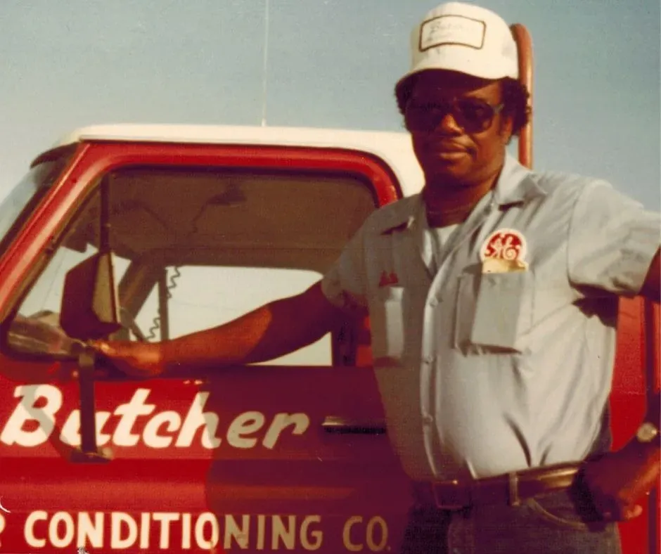 A man in uniform and cap leaning on red 'Butcher' service truck with air conditioning company signage