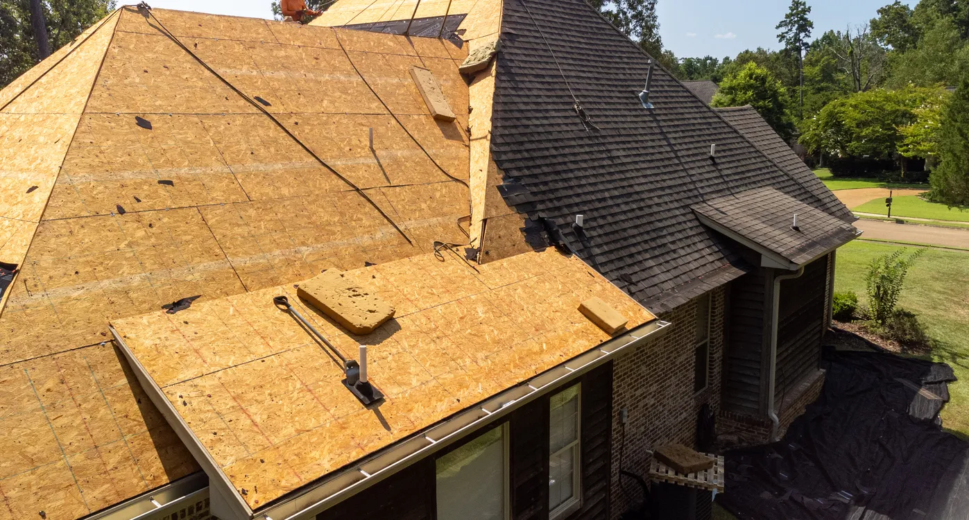 House roof under renovation with exposed plywood decking, adjacent dark shingles, roofing tools and materials visible.