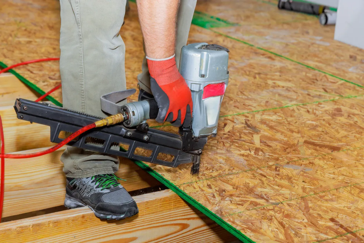 Person using pneumatic nail gun to secure oriented strand board subfloor, wearing red gloves and work shoes.