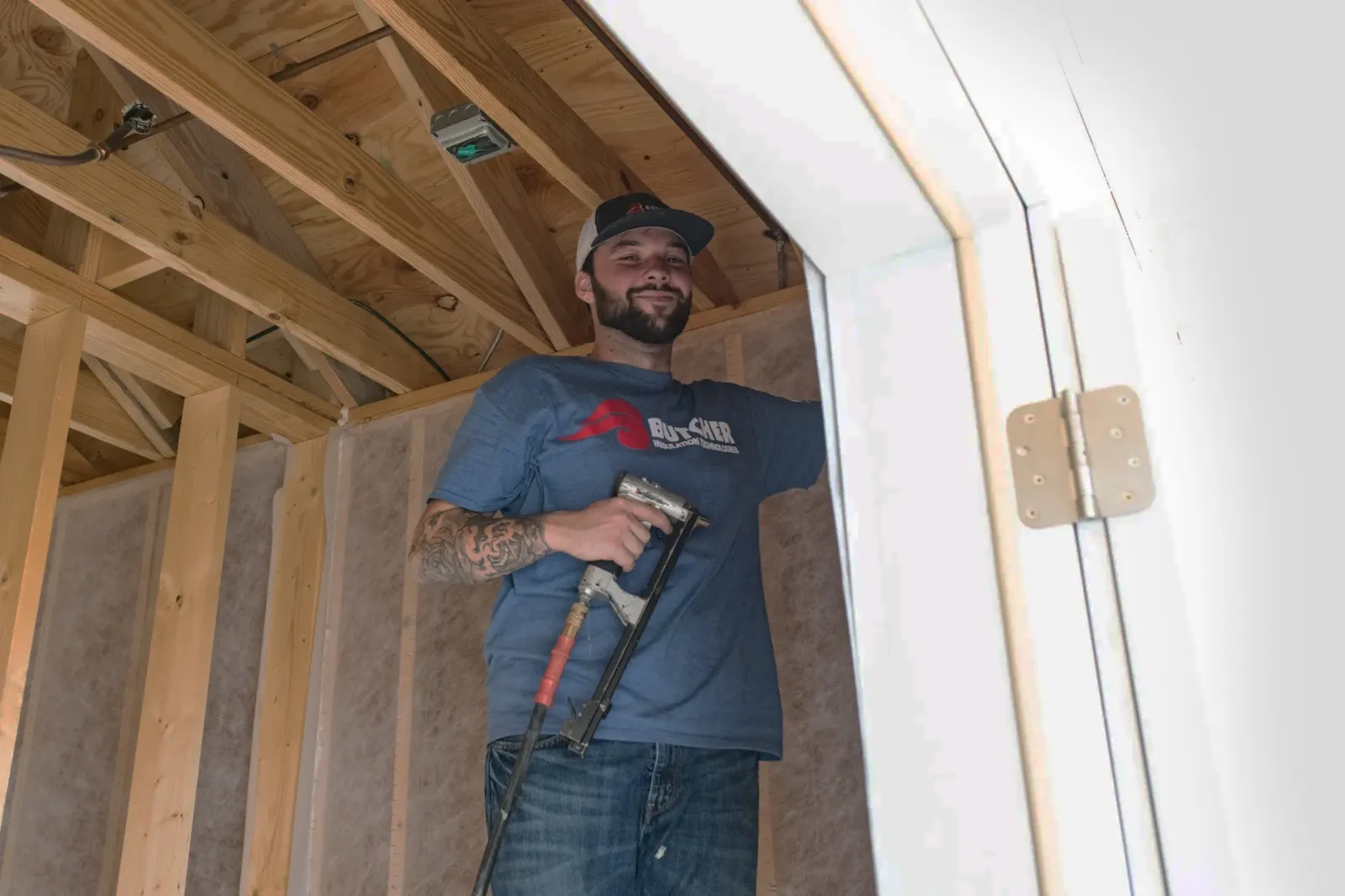 A man in a blue shirt and cap holding a nail gun while working inside a wood-framed room under construction