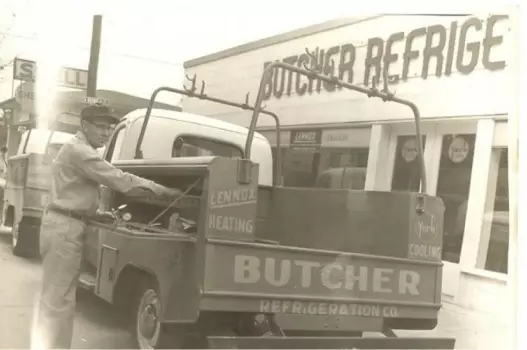 Man working at rear of vintage Butcher Refrigeration service truck parked outside company storefront.