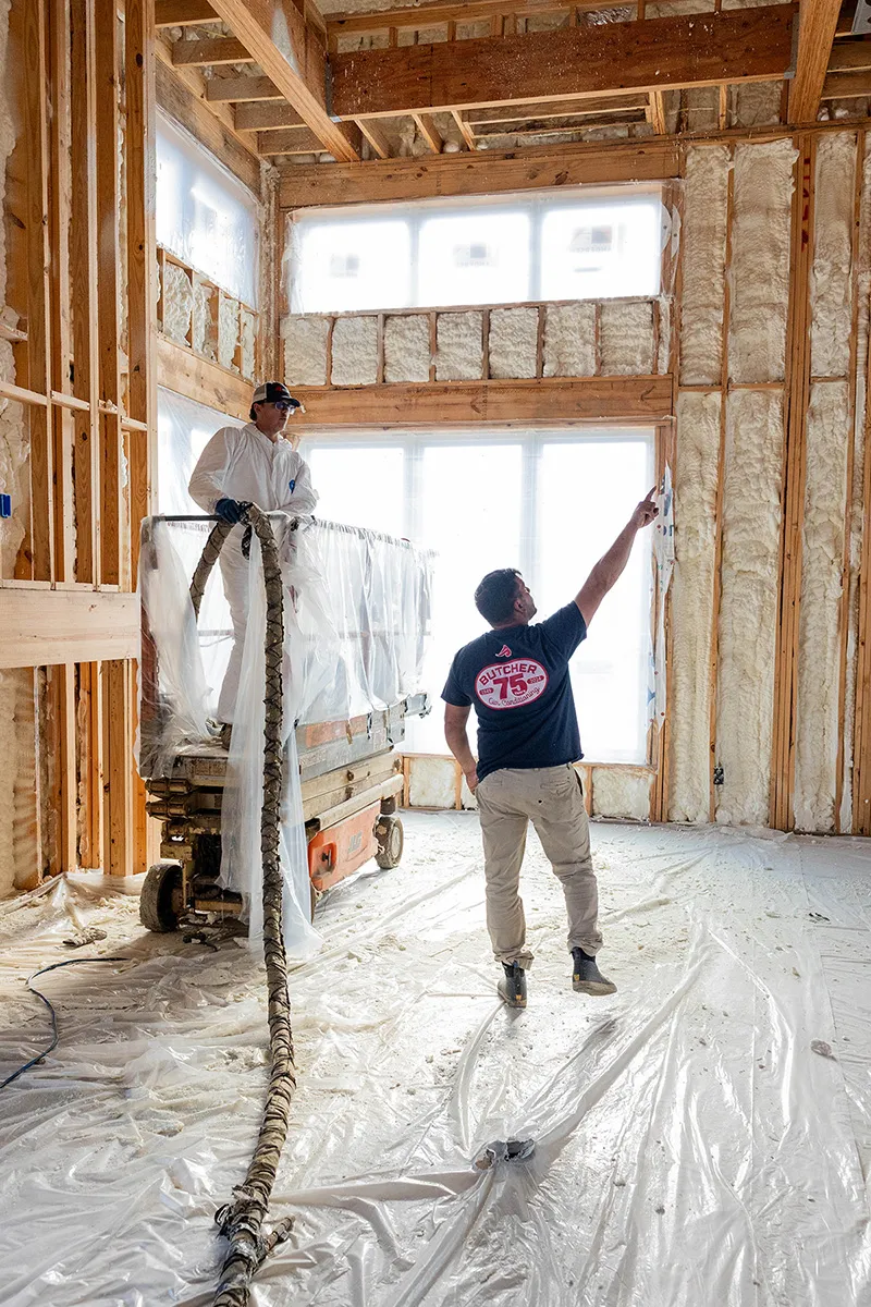 Two construction workers installing spray foam insulation in a wood-framed room, one on a lift holding hose, one pointing.